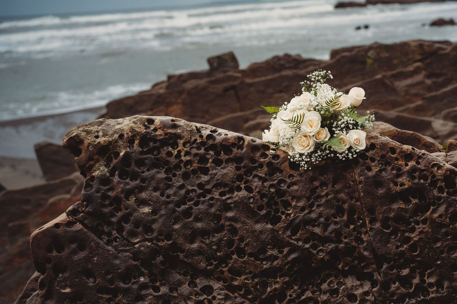 White rose wedding bouquet resting on textured volcanic rocks by the ocean shore.