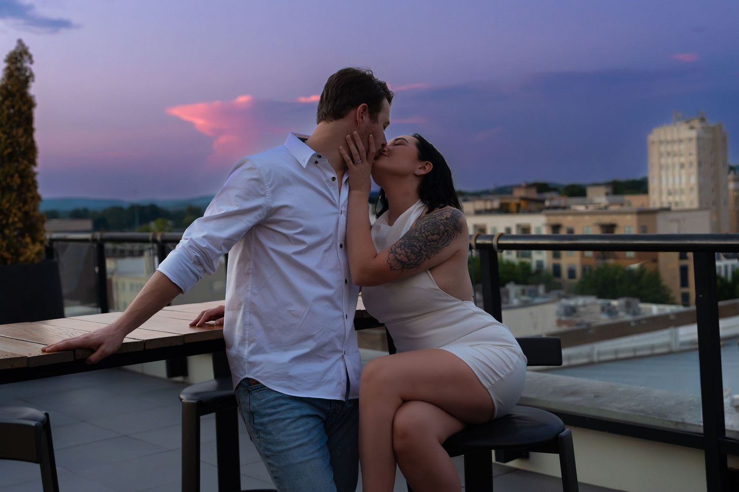 Couple kissing on a rooftop terrace at dusk, with a purple and pink sunset sky and city skyline in the background.