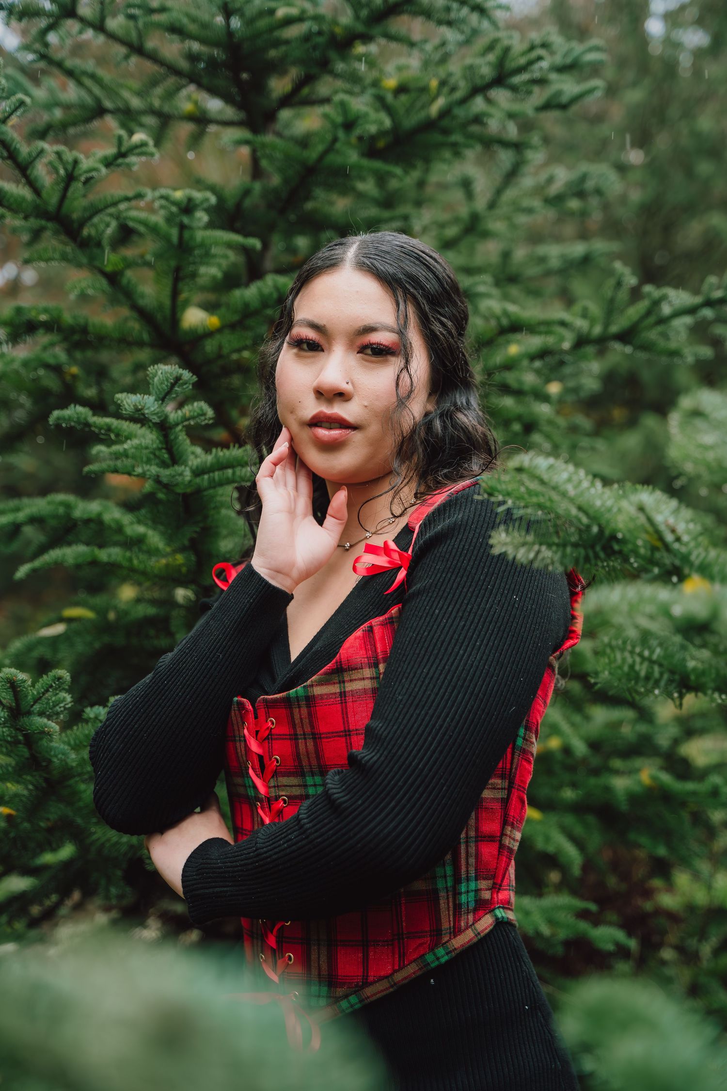 Young woman in red plaid vest and black sweater poses among green pine trees in a festive outdoor setting.