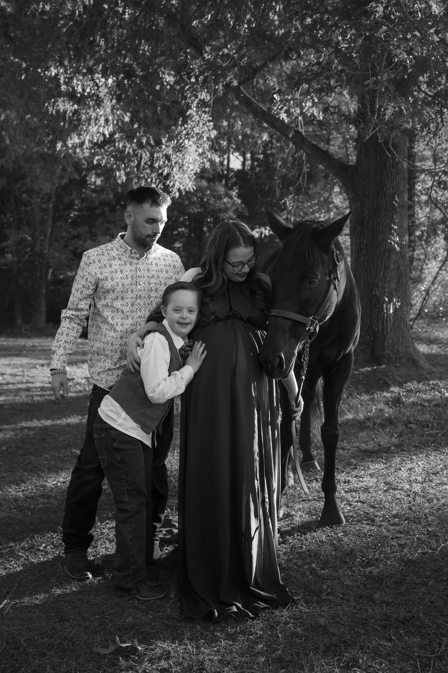 Black and white family photo with pregnant woman, man, child, and horse under trees outdoors.