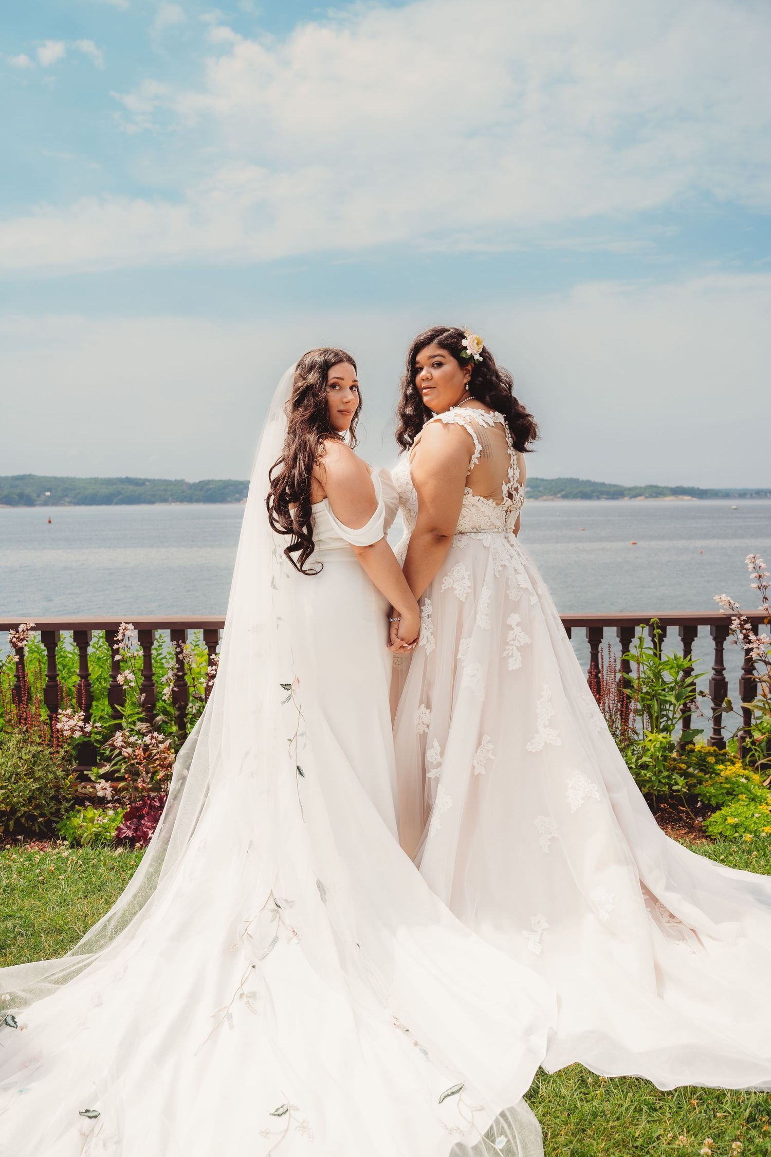 Two brides in white wedding gowns holding hands overlooking scenic waterfront. Massachusetts