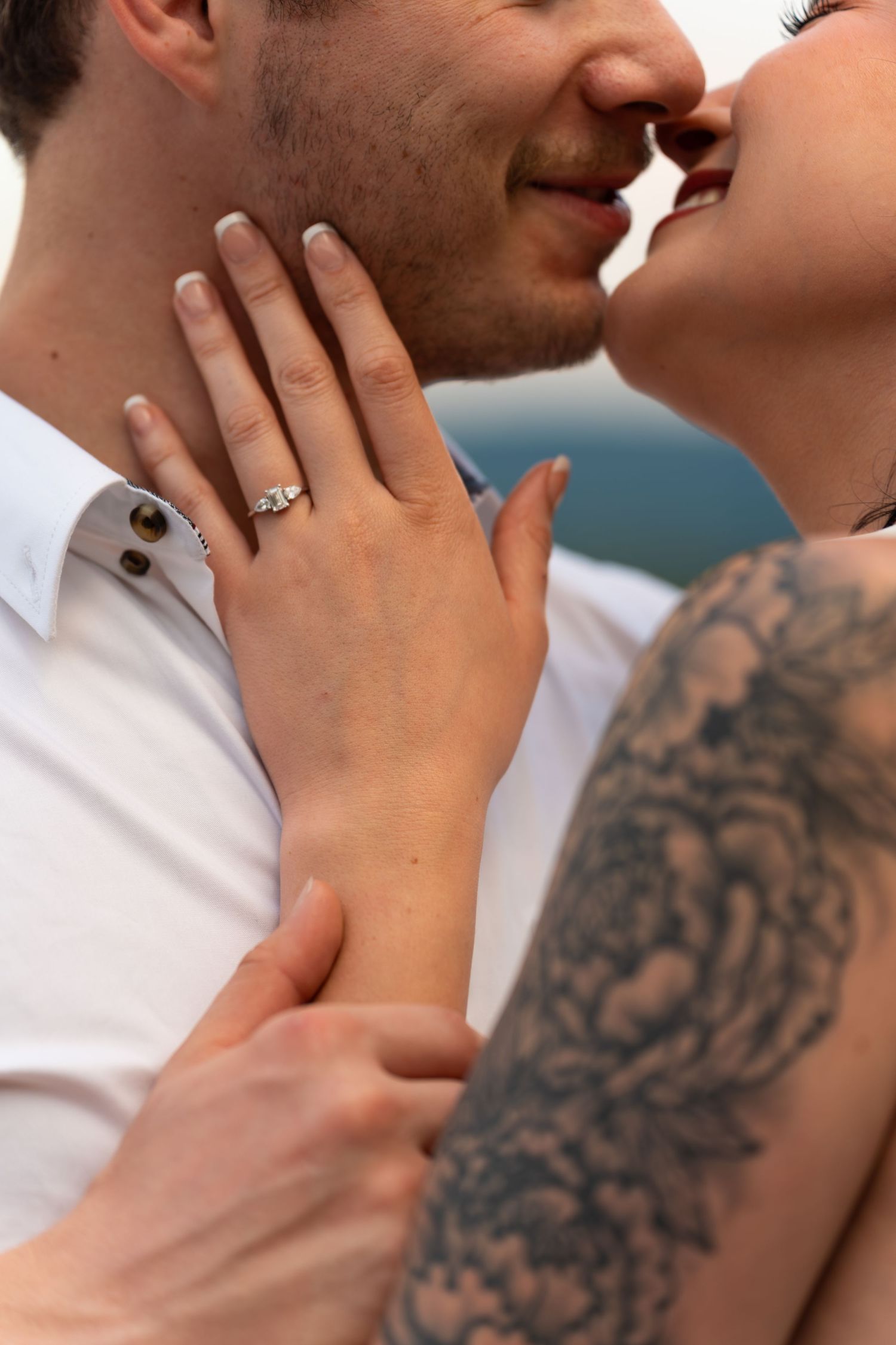 Couple sharing a romantic kiss, woman displaying diamond engagement ring and floral tattoo on arm.