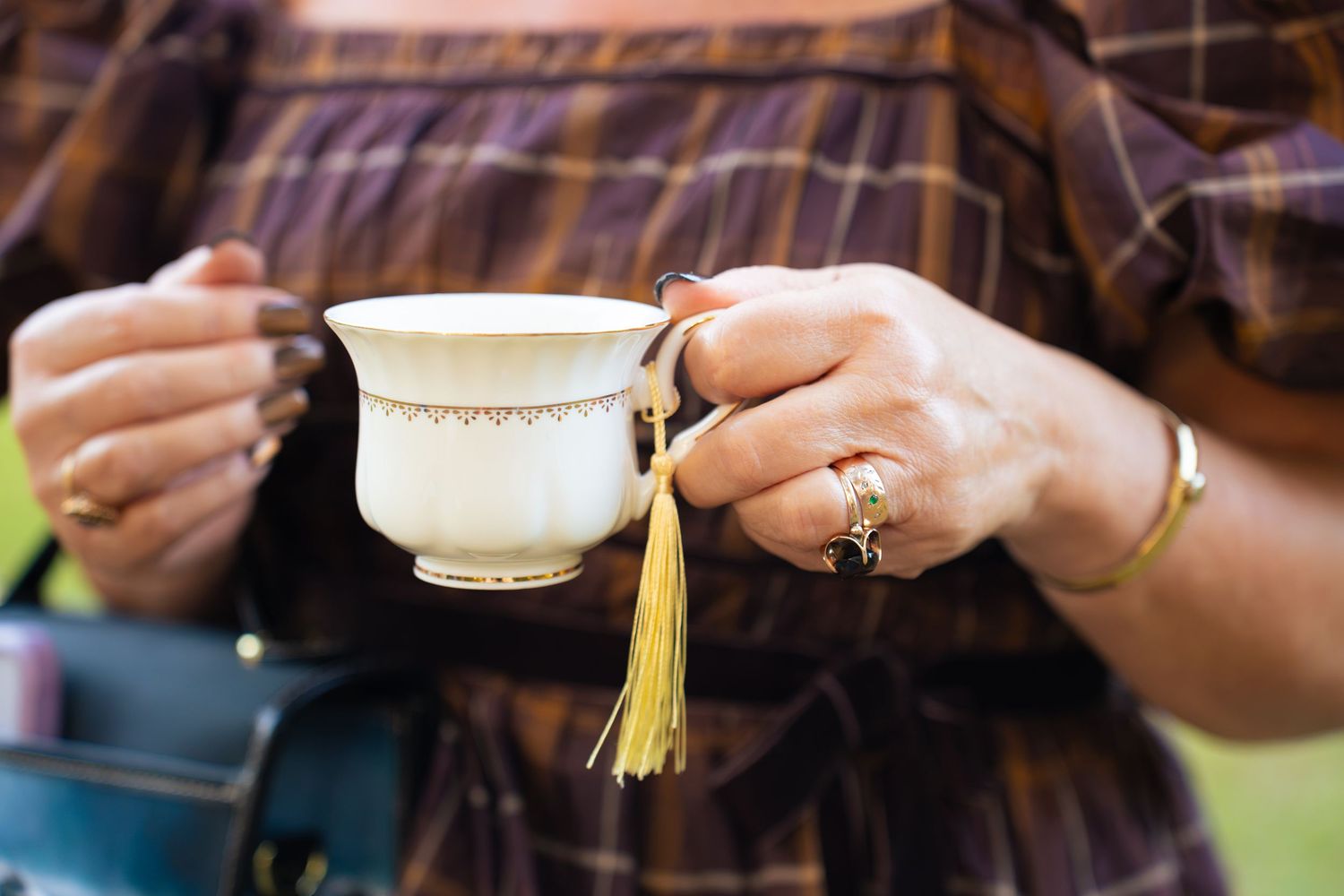 Woman's hands holding a white porcelain teacup with gold trim and a golden tassel, wearing rings and a bracelet with a plaid dress.