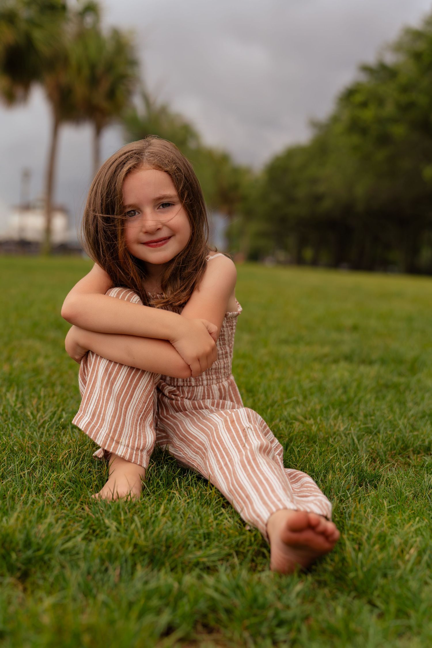 A young girl in striped overalls sits barefoot on green grass, smiling with arms crossed over her knees.