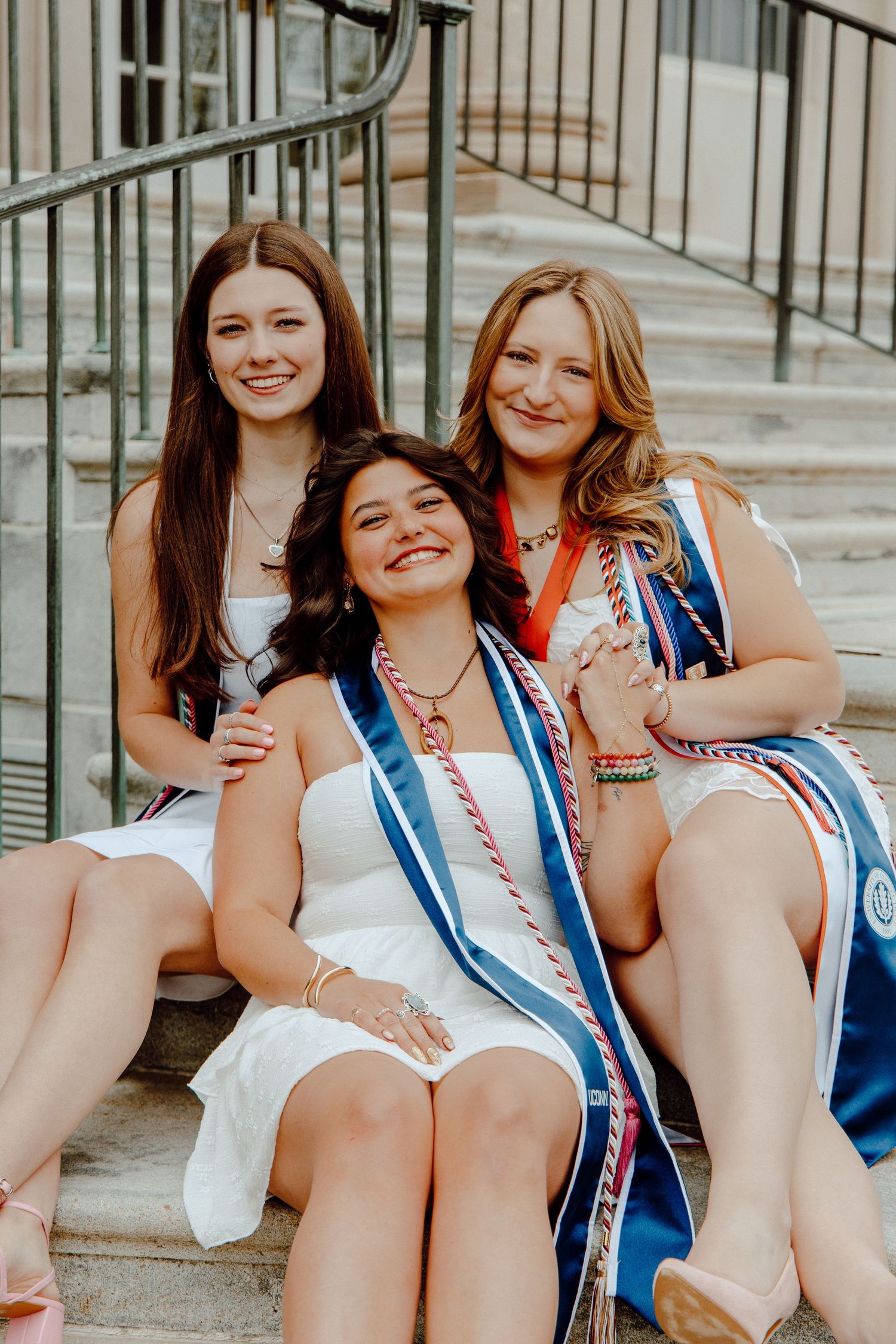 UConn graduation session featuring three seniors in white dresses and stoles, celebrating achievement with bright smiles on campus steps.