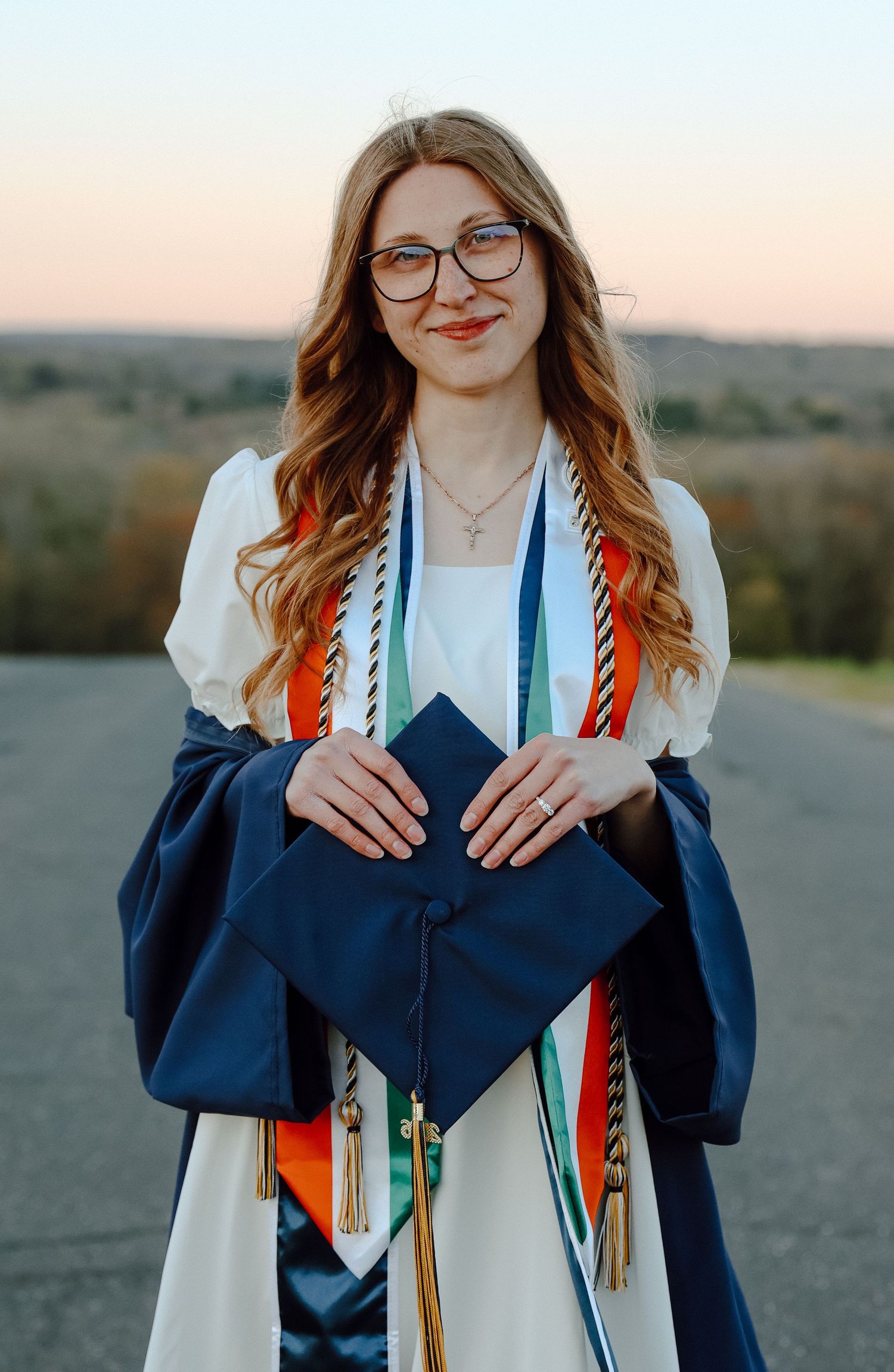 UConn senior on Horsebarn Hill at sunset, celebrating graduation with honor cords, Husky pride, and glowing golden-hour scenery.