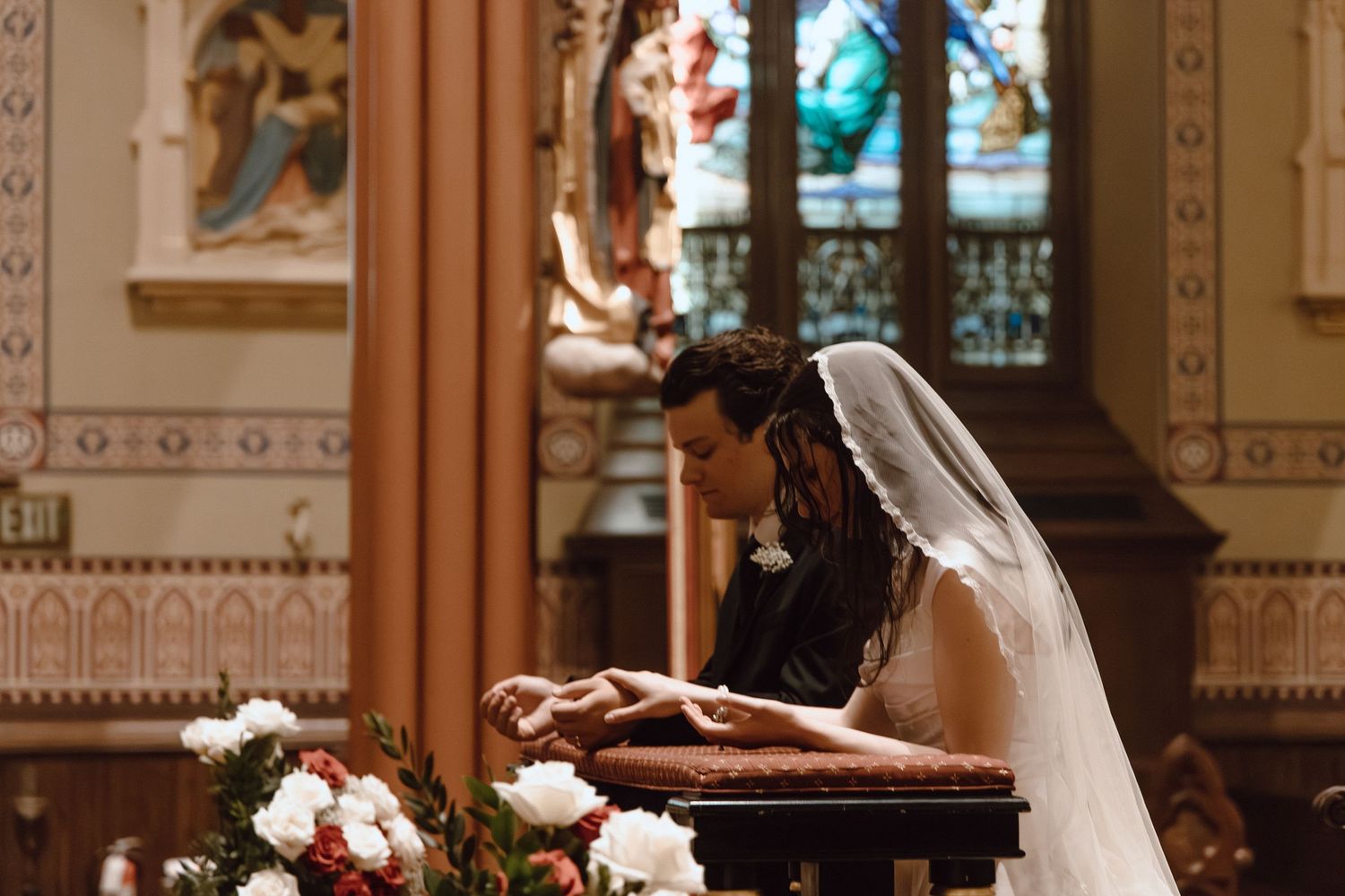 Catholic wedding moment at the altar, bride and groom kneeling in prayer surrounded by ornate church details and sacred beauty.