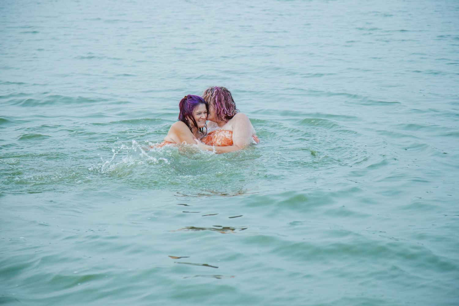 Two people splash and play together in calm blue waters near the shoreline.