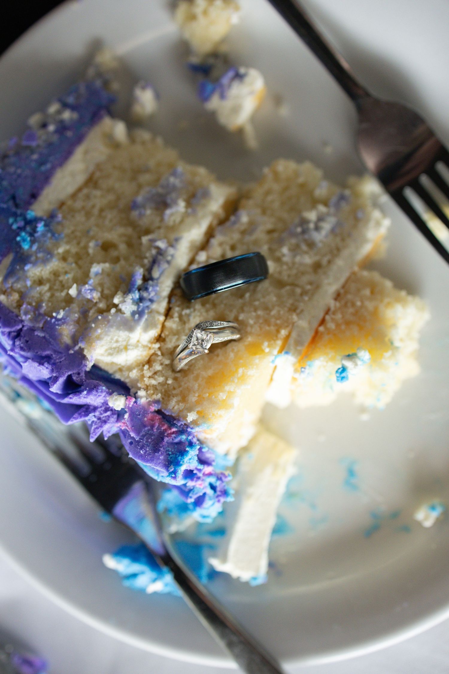Wedding rings resting on a slice of purple and white layered cake on a white plate with forks.