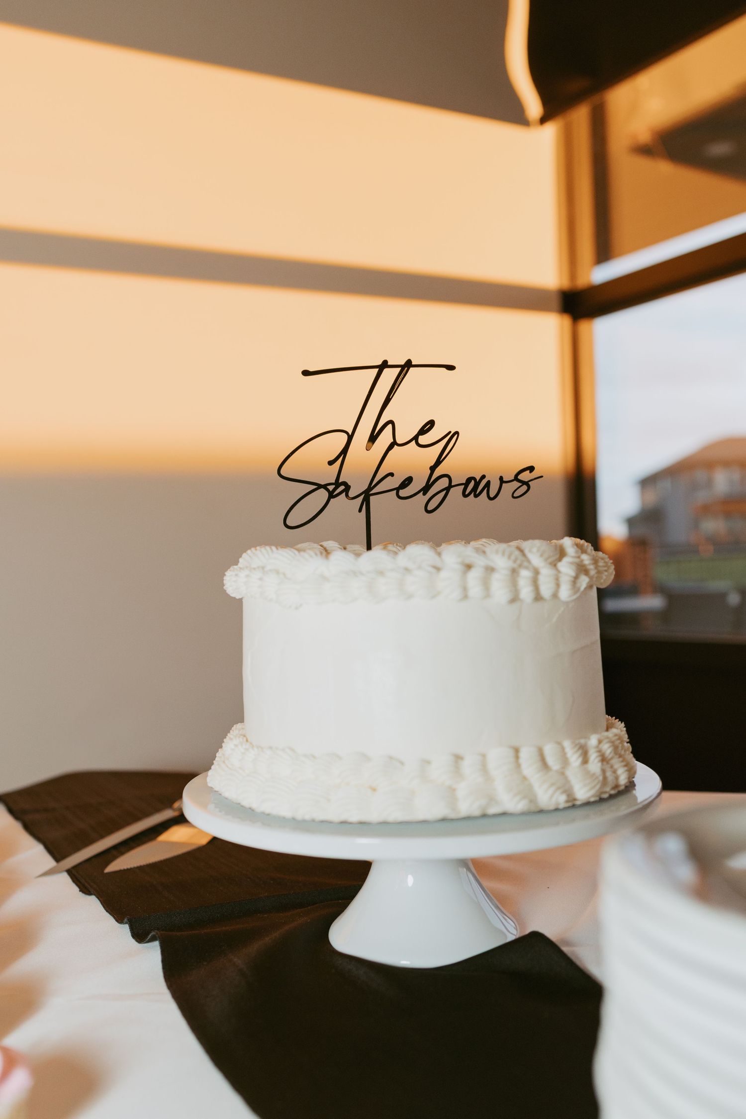 White wedding cake with 'The Sakebows' cake topper on a white stand, golden sunset light in background.