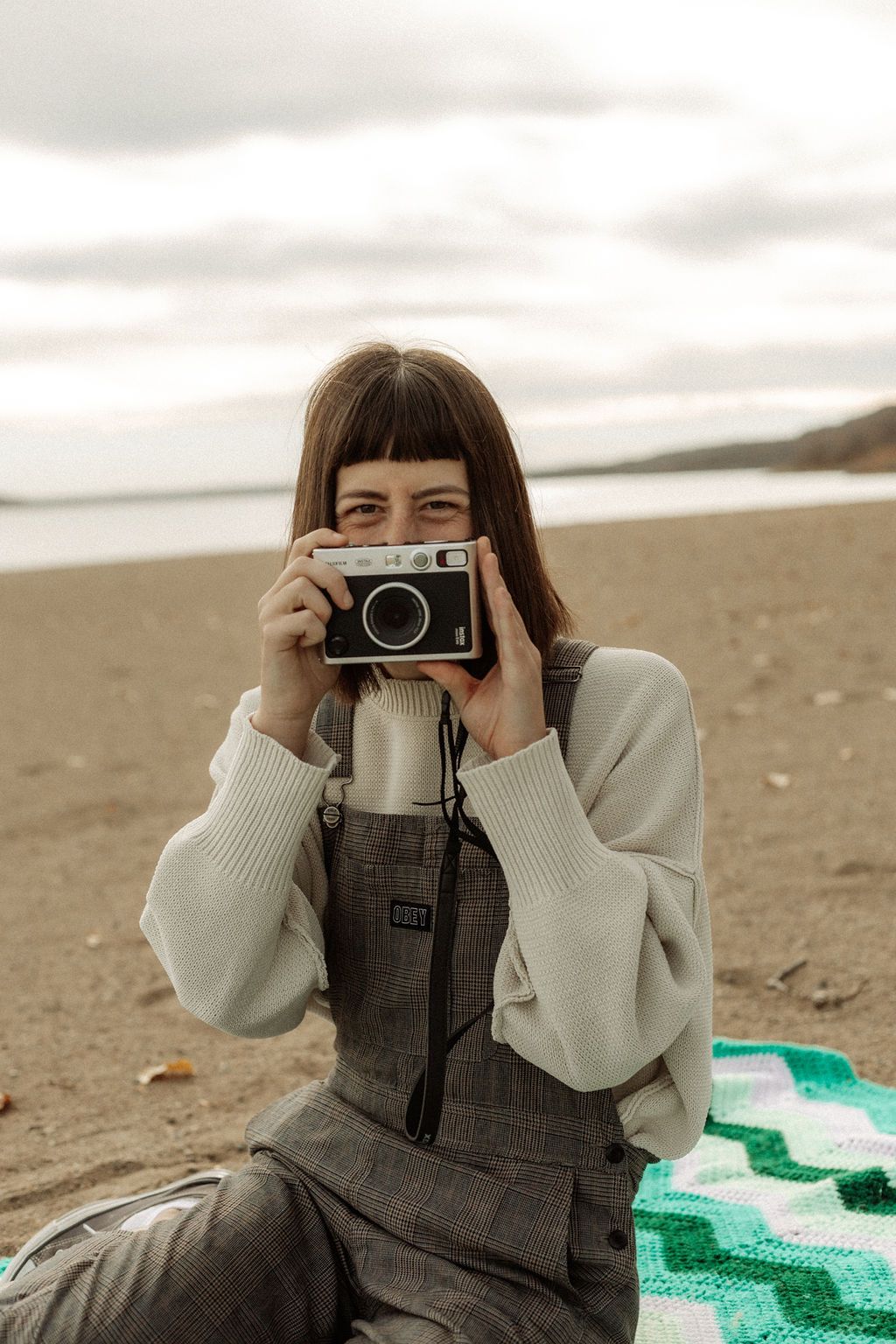 Photographer in gray sweater and overalls takes pictures with vintage camera while sitting on beach blanket.