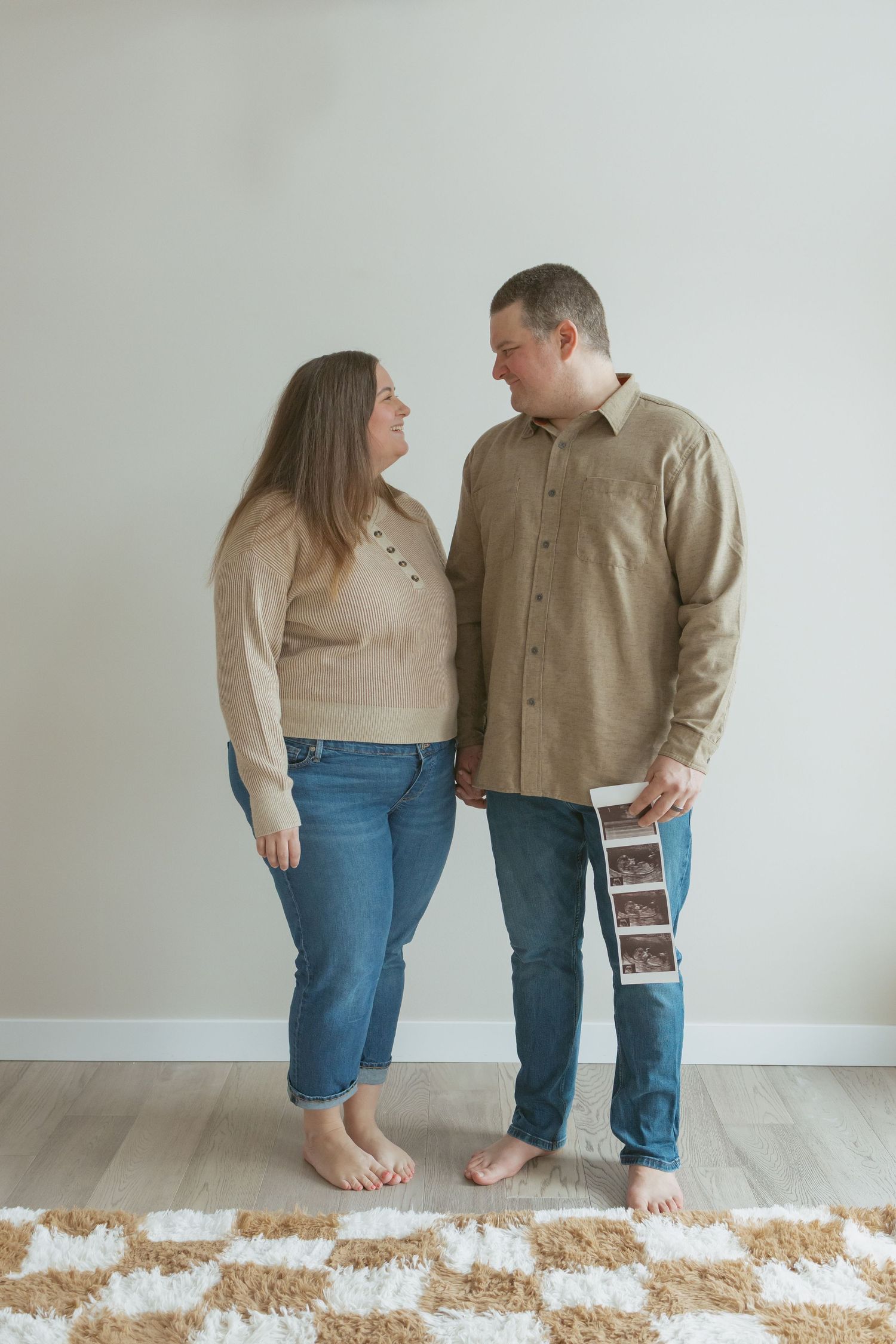 Couple holding hands and ultrasound photos, smiling at each other in a cozy home setting, announcing pregnancy.
