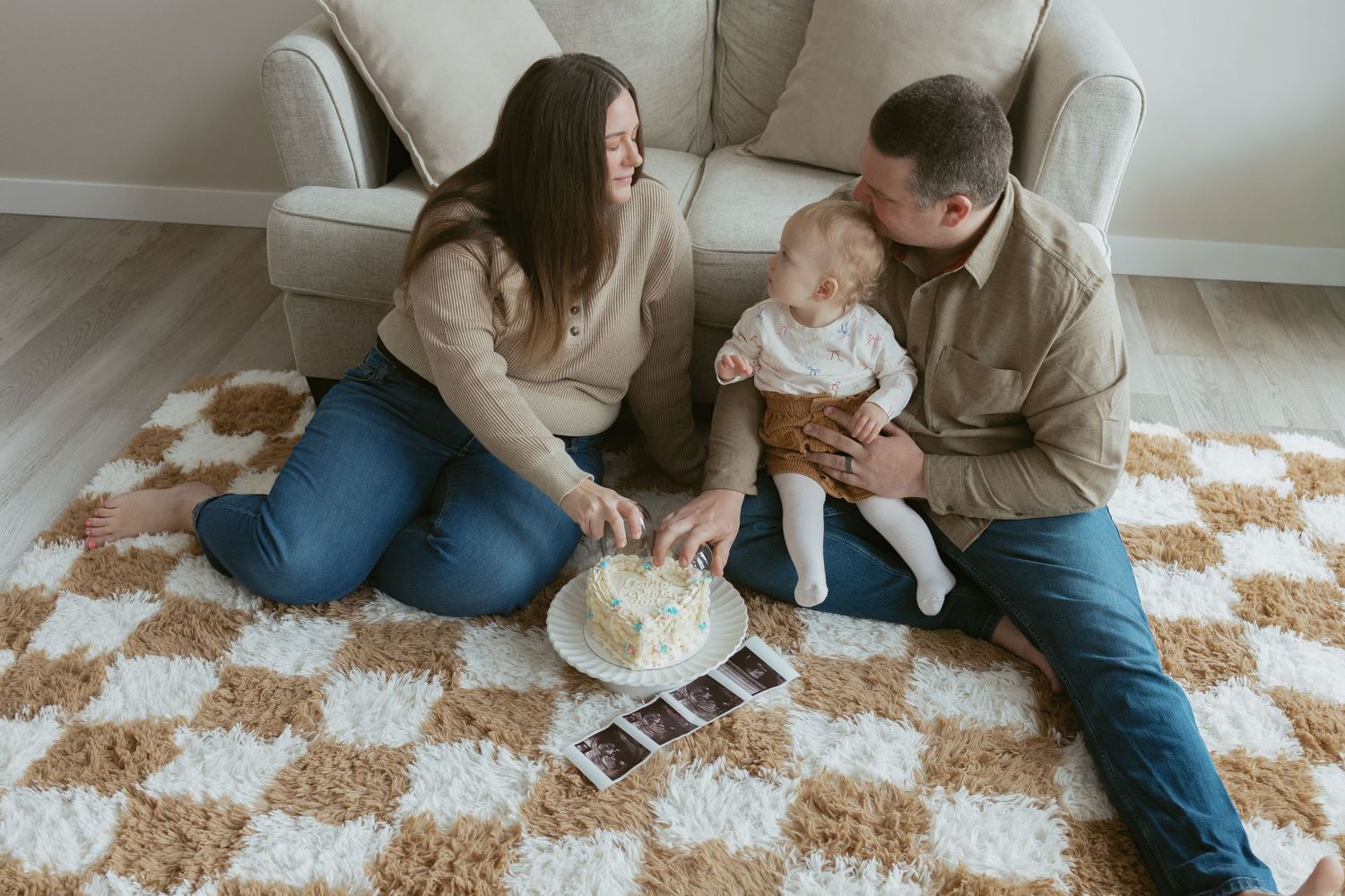 Family sitting on a checkered rug celebrating baby's first birthday with a small cake and ultrasound photos nearby.