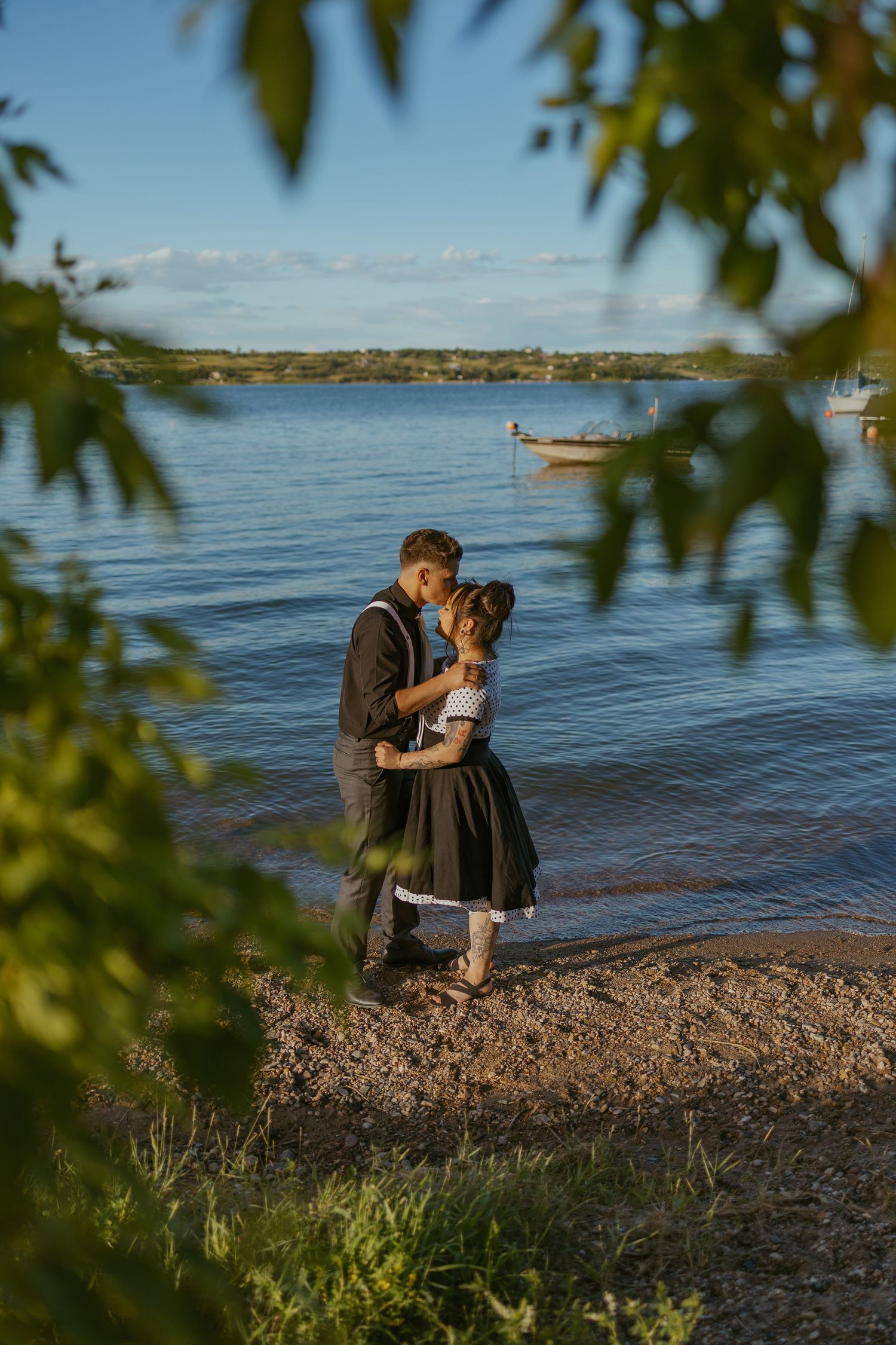 Romantic couple sharing intimate moment on rocky shoreline framed by tree leaves.