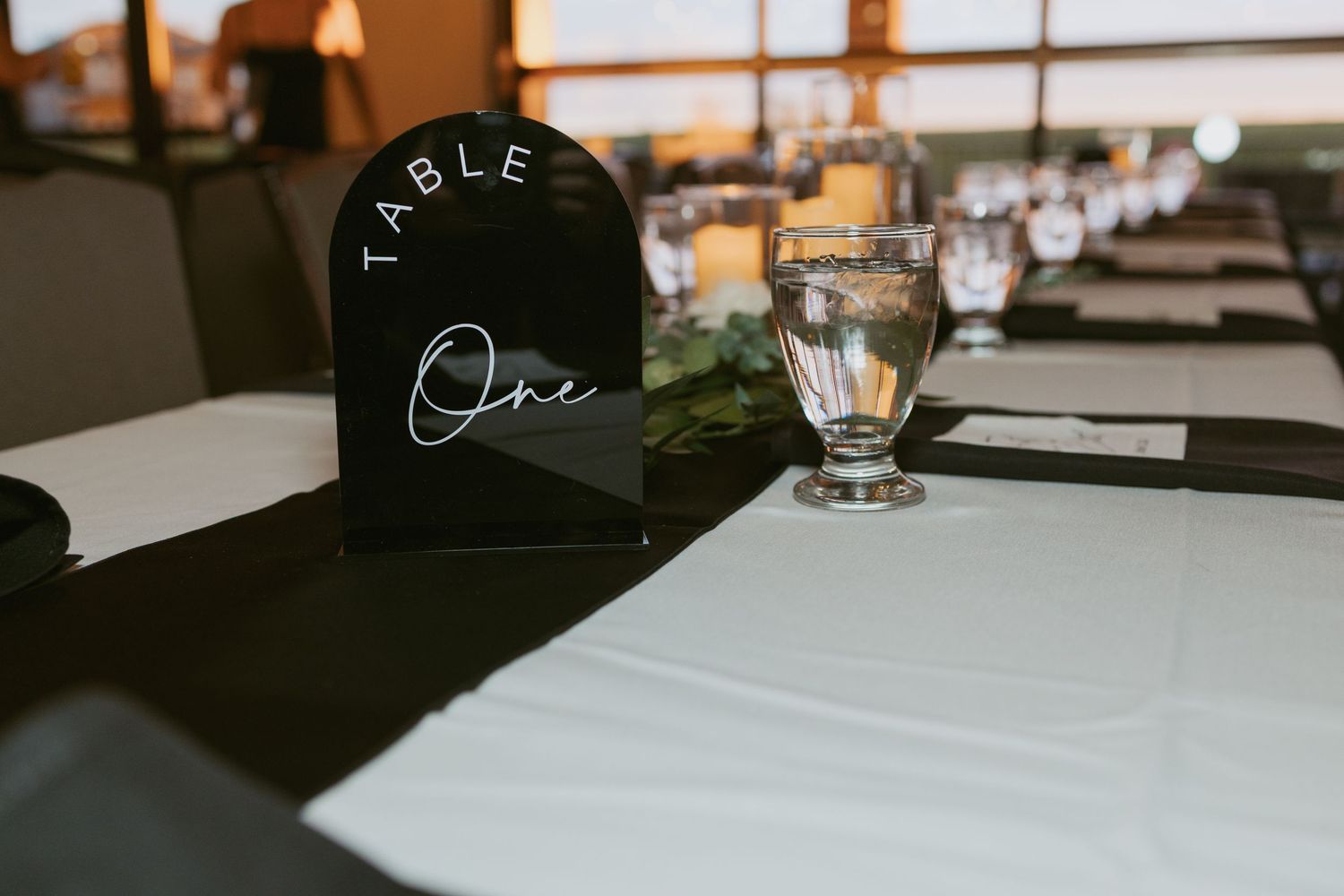 Elegant wedding reception table setup with black tablecloth, Table One acrylic sign, and crystal glassware in a dimly lit venue.
