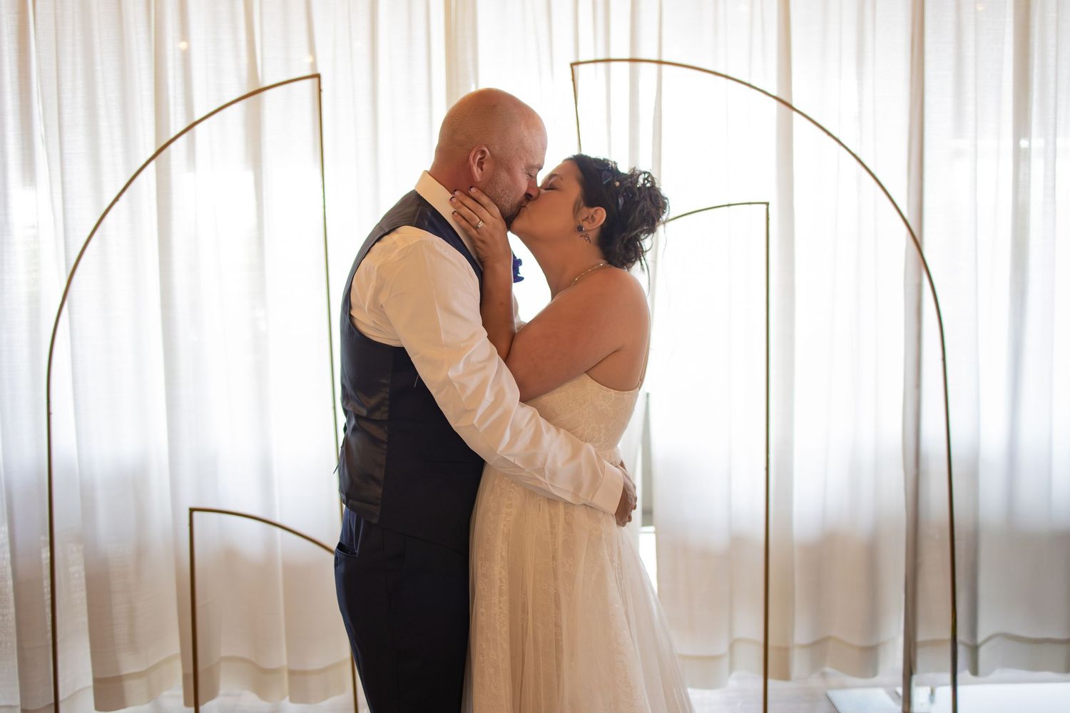 Bride and groom share first kiss under gold arch with sheer white curtains backdrop at wedding ceremony.