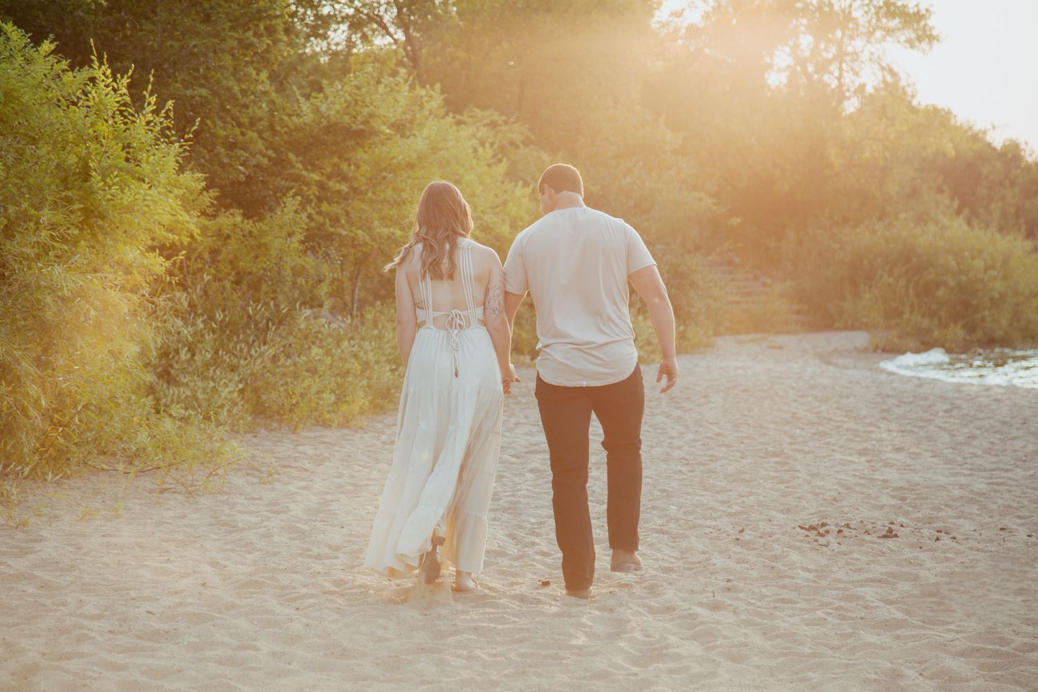A couple walks hand in hand on a sandy beach path at sunset.