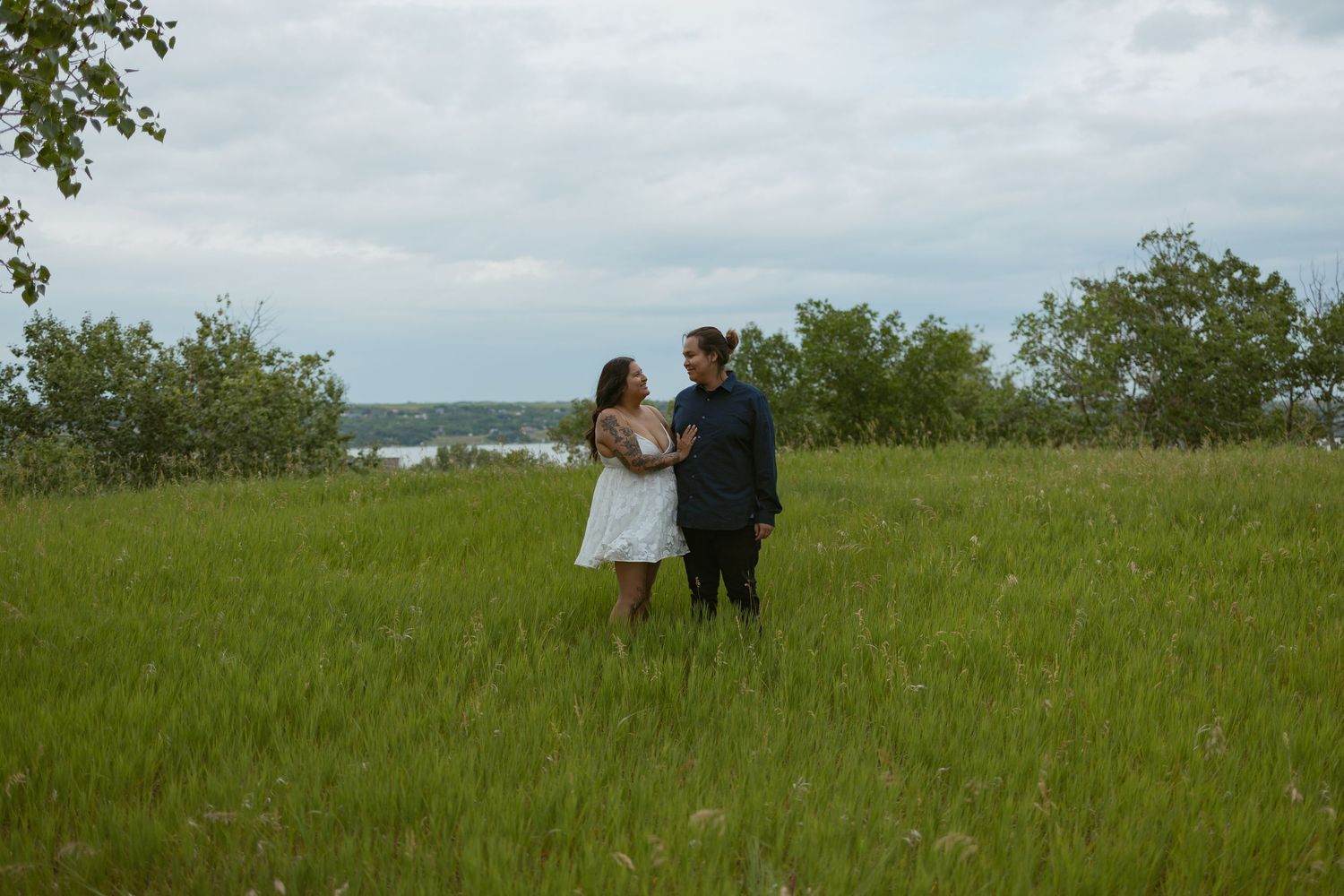 A couple stands together in a grassy meadow with trees and water in the background on an overcast day.
