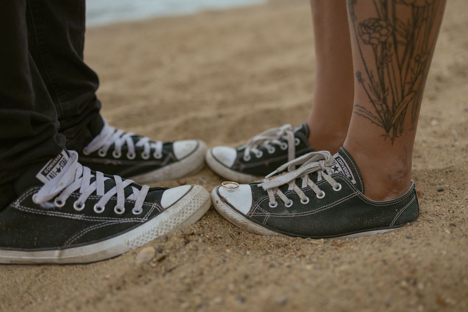 Two pairs of worn Converse sneakers standing toe to toe on sandy ground.