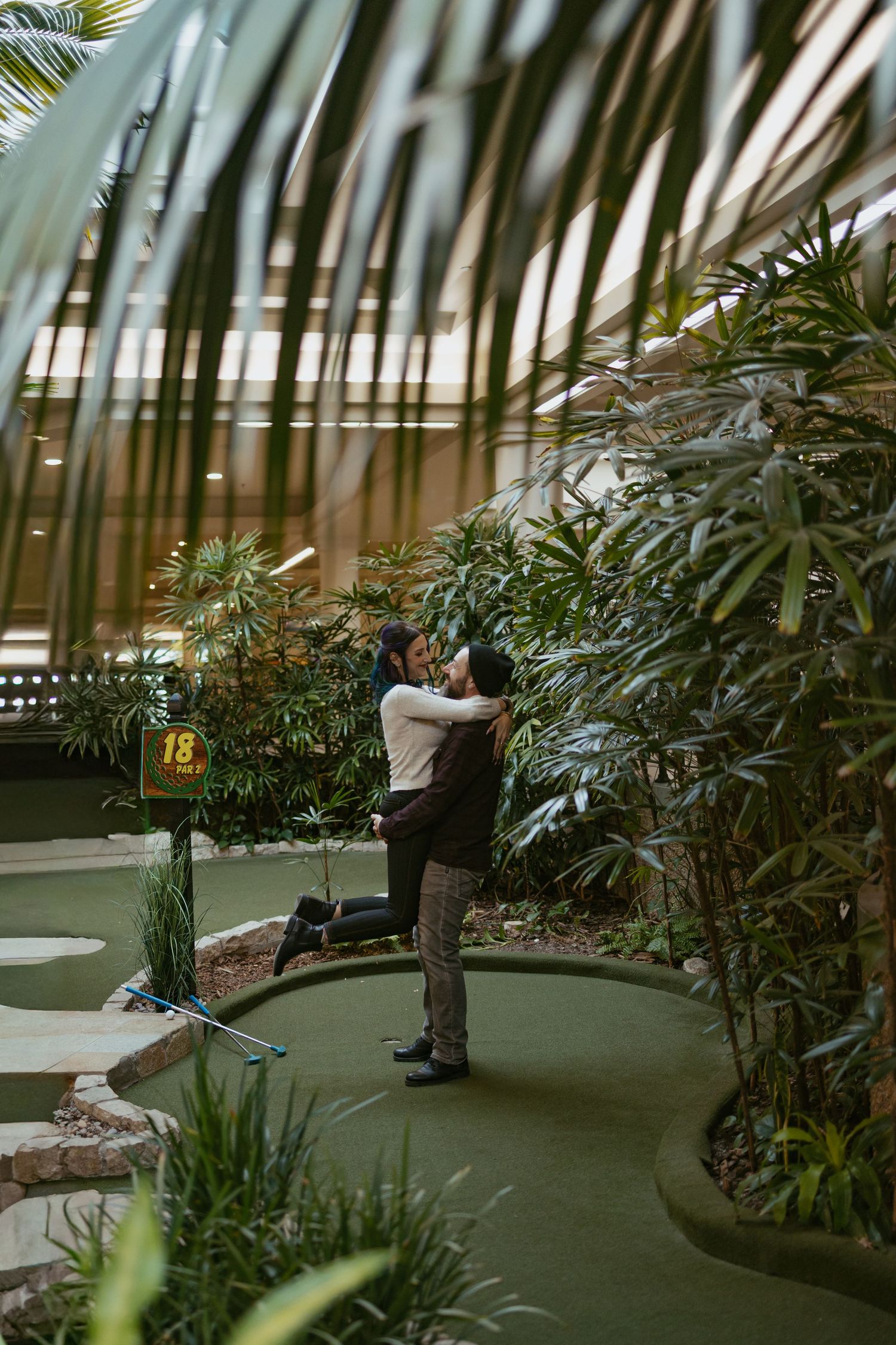 Couple embracing on hole 18 of an indoor mini golf course surrounded by lush tropical plants.