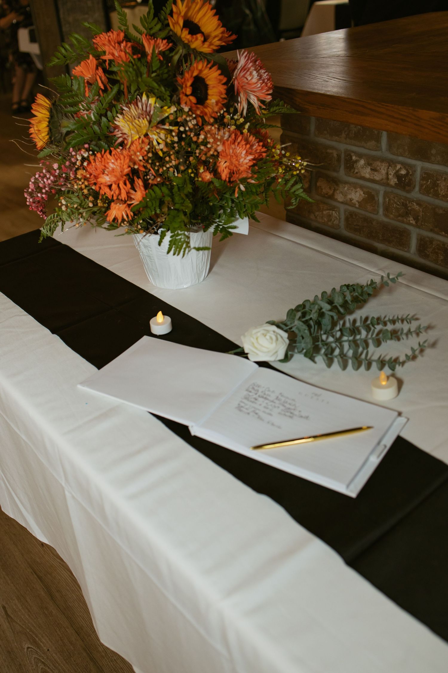Elegant wedding signing table with orange floral arrangement, guest book, gold pen, white rose, and candles on black runner.