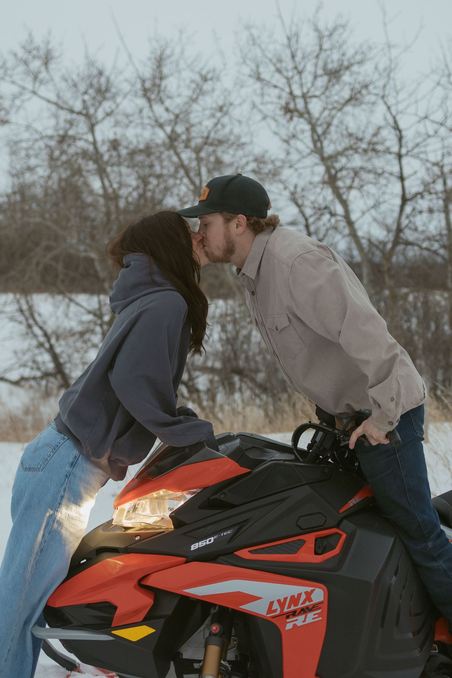 Couple sharing a romantic kiss on a red Lynx snowmobile in a snowy winter landscape.