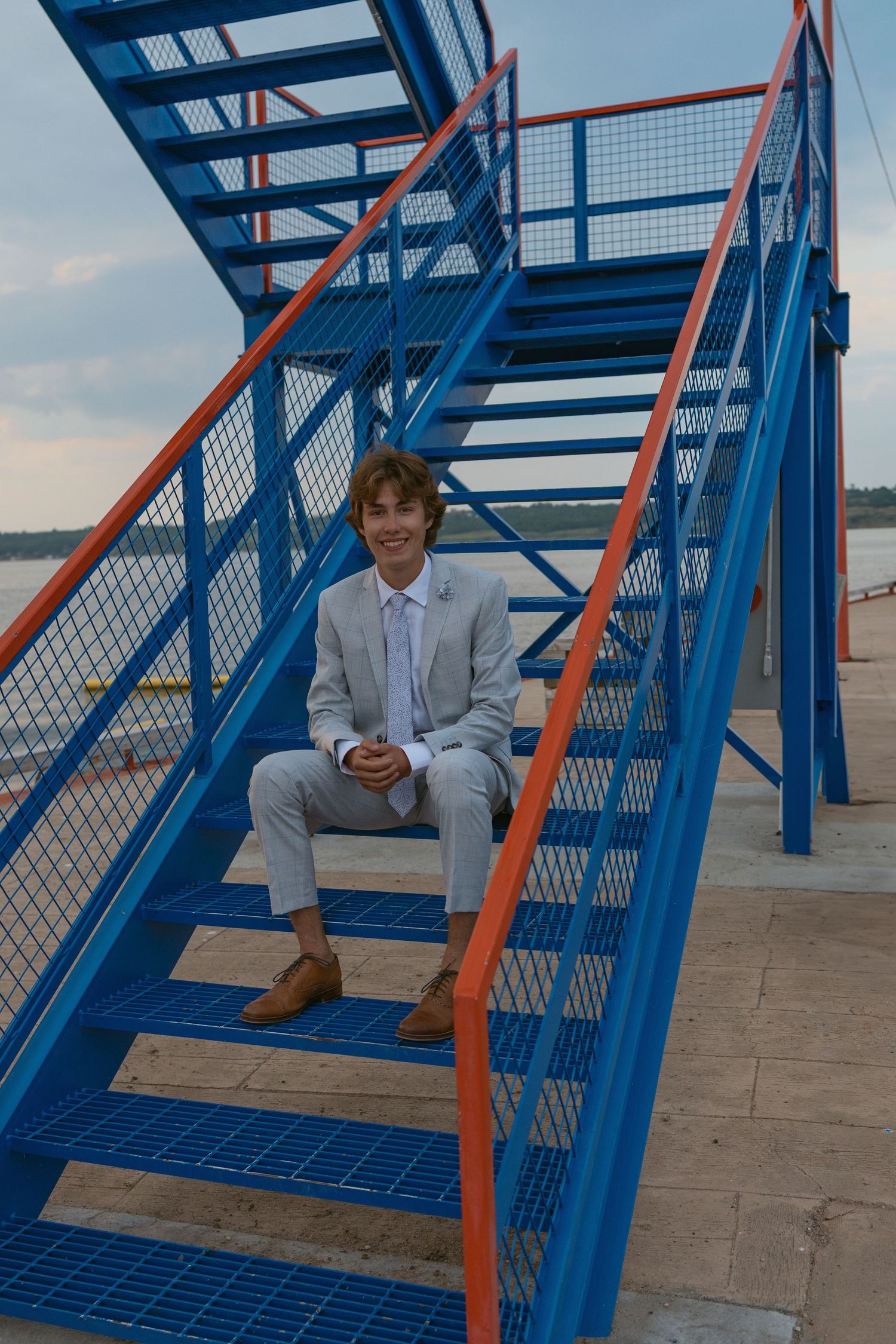 Person in light suit sits on blue metal stairs outdoors near waterfront.