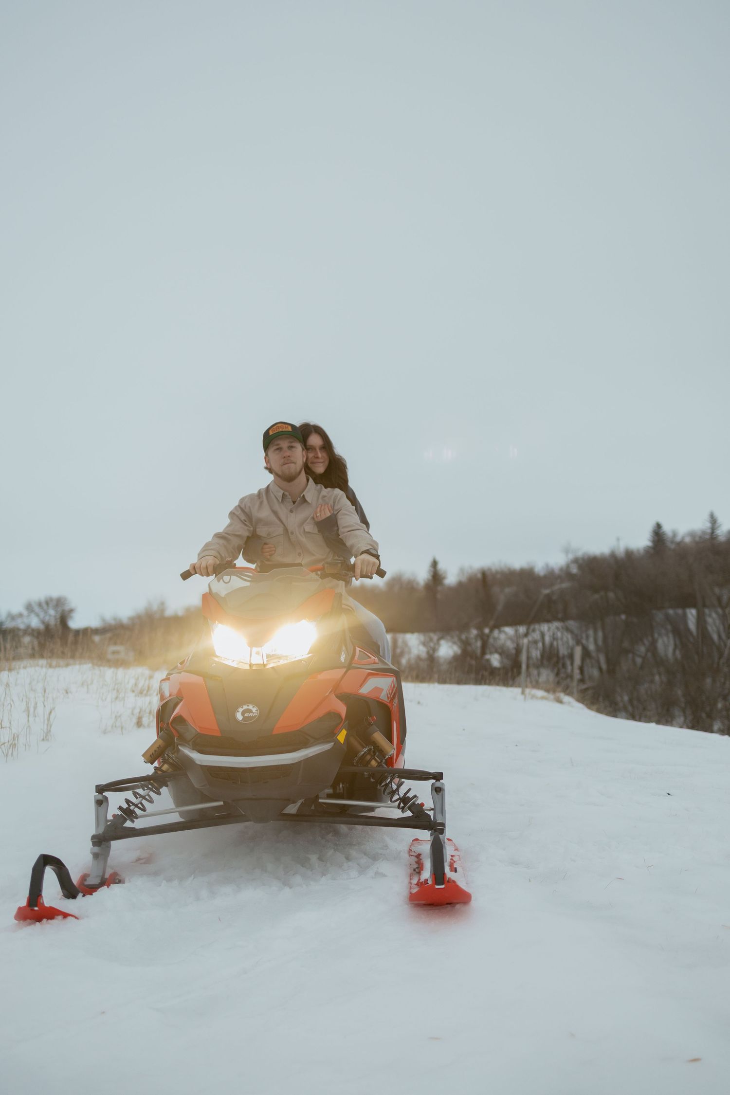 Couple riding a red snowmobile together on a snowy winter landscape with bright headlights glowing.