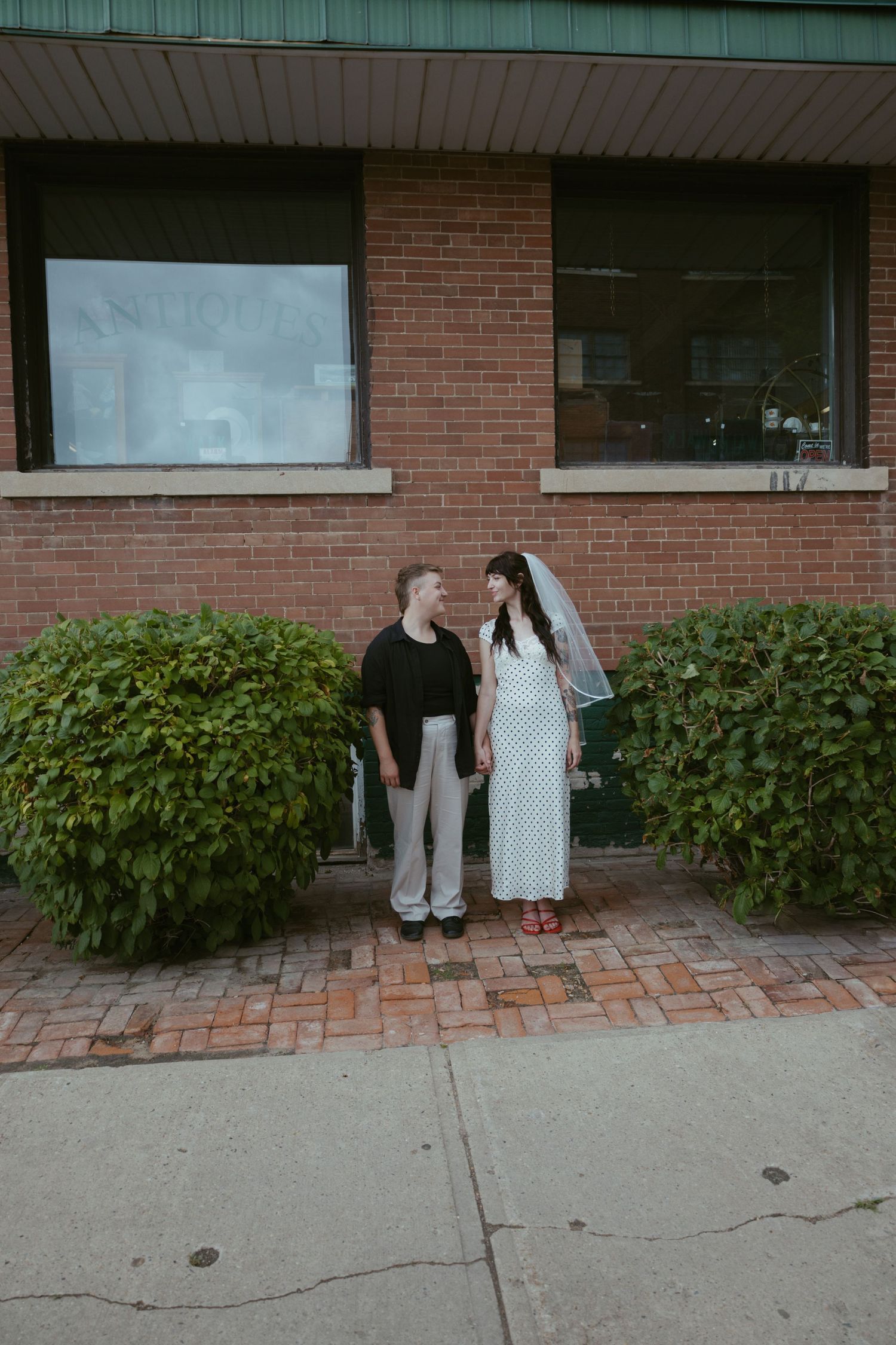 A couple standing by a brick building with landscaped hedges.