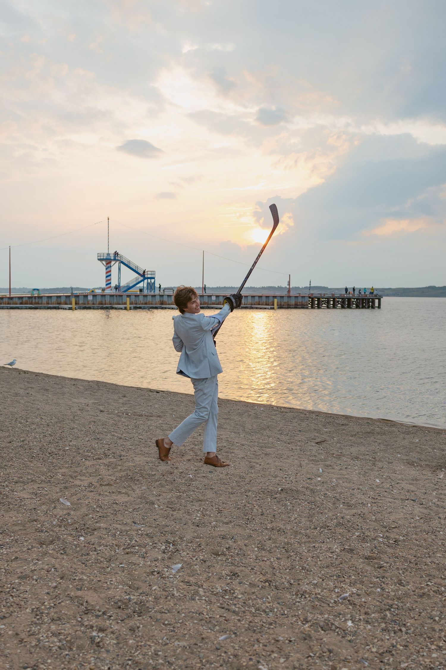 A person in a light colored suit throws stones on a beach at sunset with a pier visible in the background.