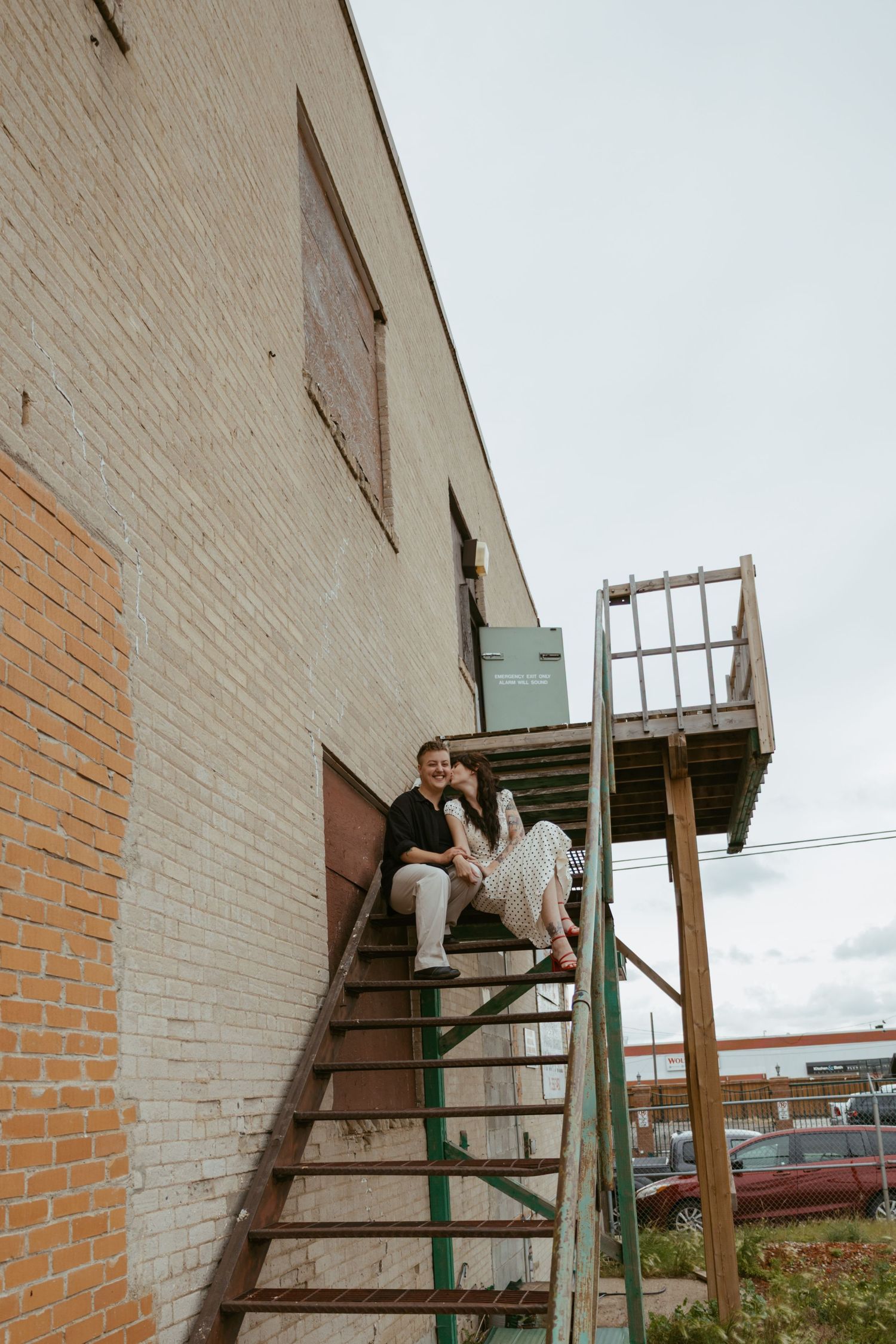 Two people sitting on exterior metal stairs against a brick building wall.