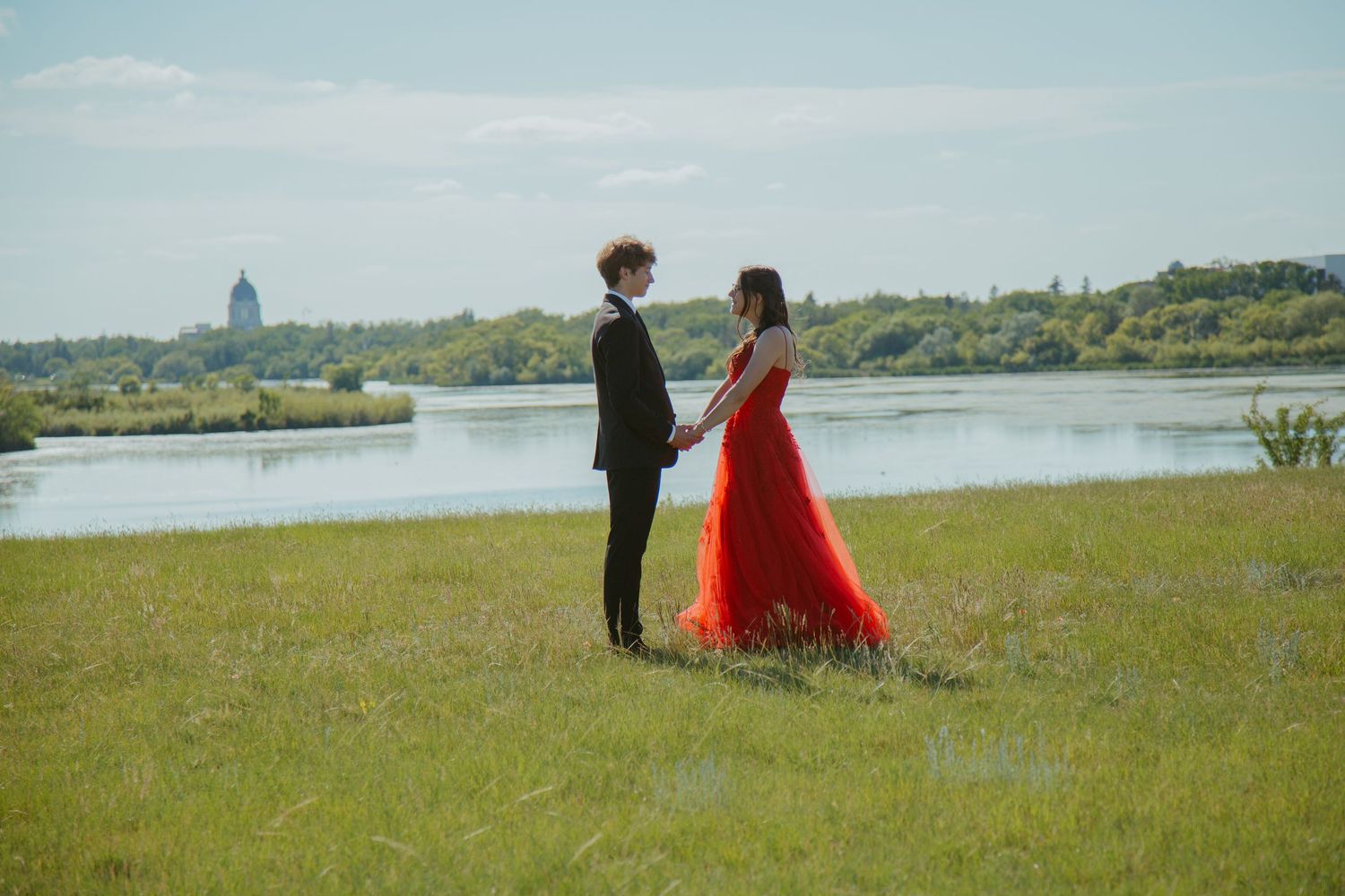 Couple standing in grassy field overlooking waterfront on sunny day.