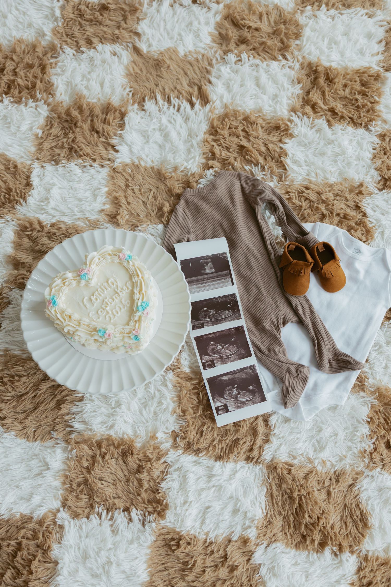 Heart-shaped baby announcement cake beside ultrasound photos, tiny onesie, and brown moccasins on a checkered fluffy rug.