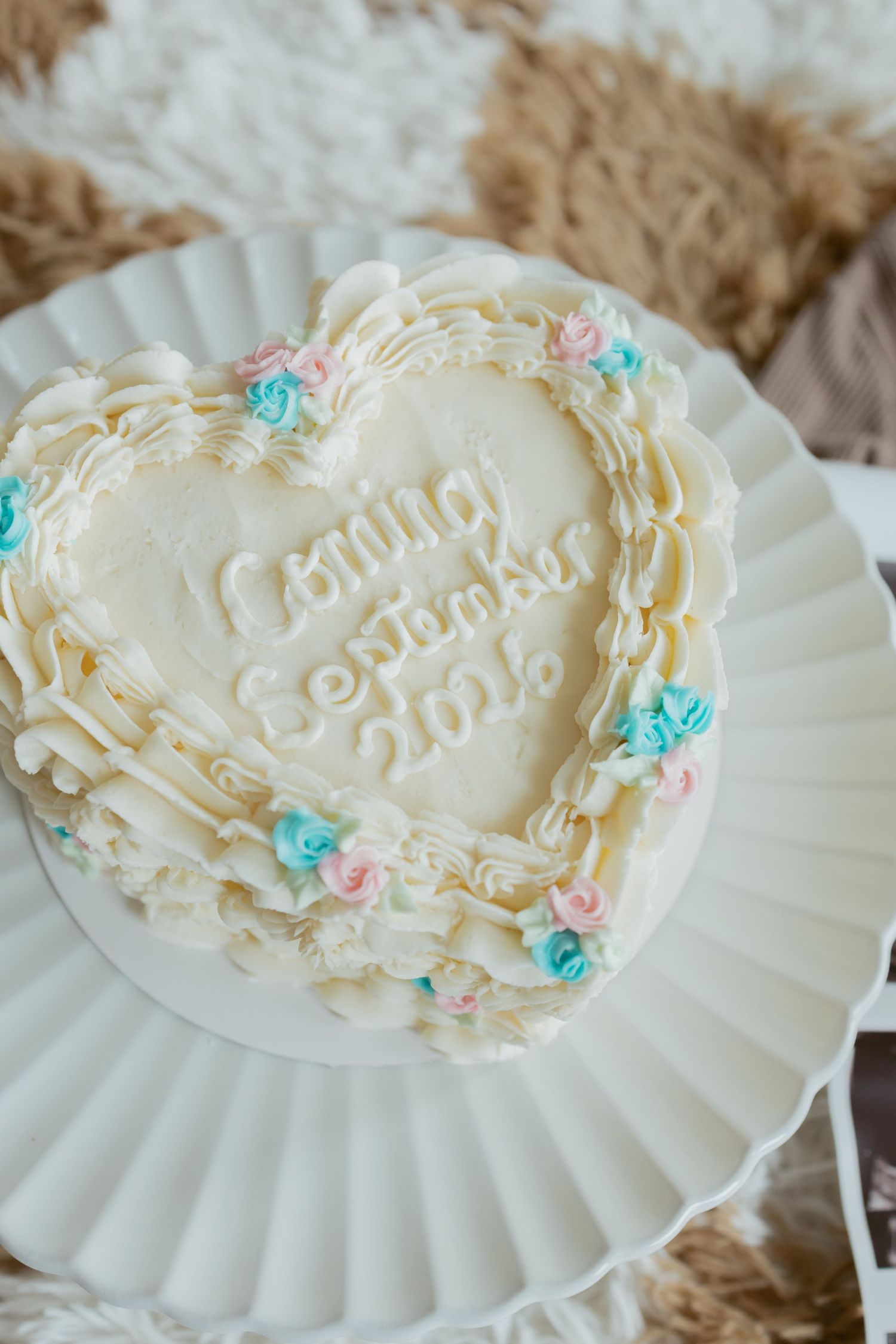 Heart-shaped white frosted baby shower cake with pink and blue roses reading Coming September 2016 on white cake stand.