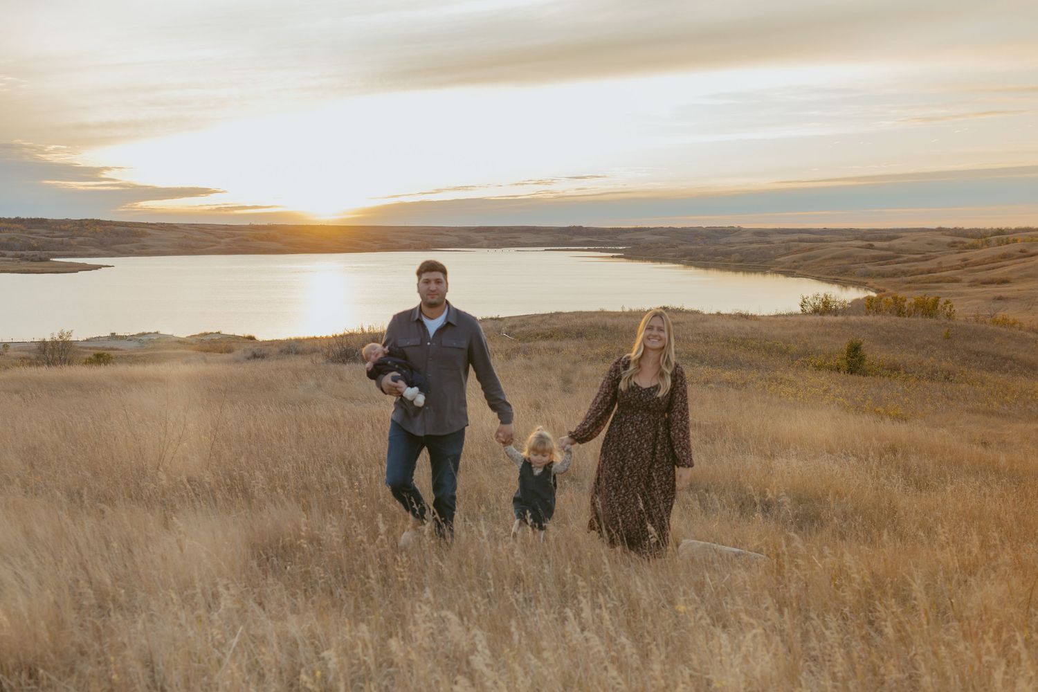 Family of three walking through golden prairie grass at sunset with a lake and glowing sky in the background.