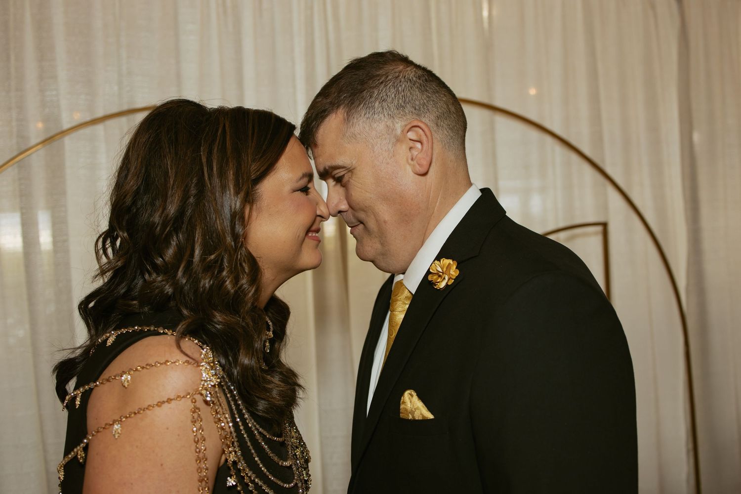 Couple in formal attire sharing an intimate moment, man in black suit with gold tie, woman in elegant gown, circular arch backdrop.