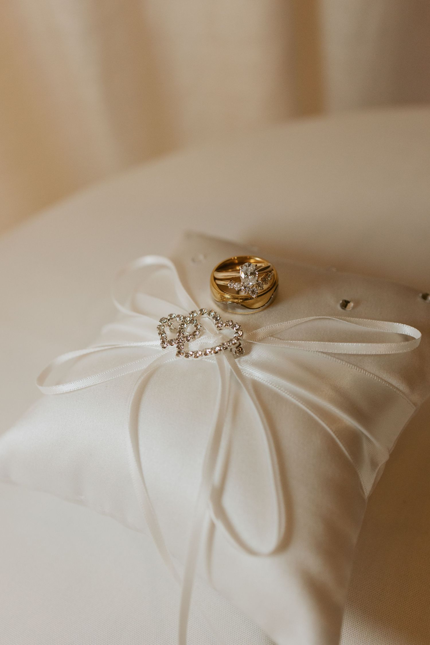 Gold diamond engagement ring and crystal brooch displayed on white satin ring bearer pillow with ribbon bow.