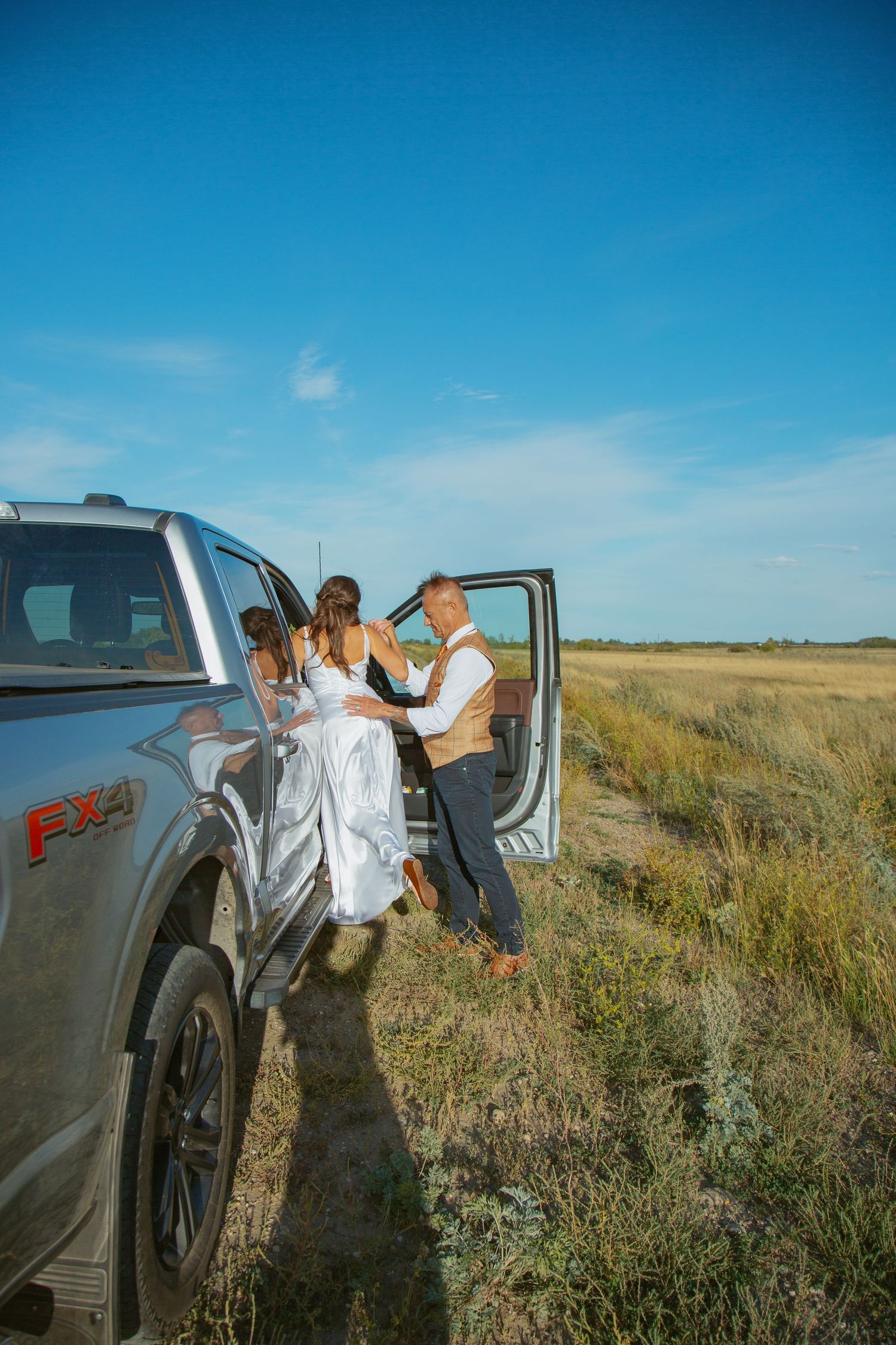 Couple by pickup truck in rural field during wedding photoshoot.