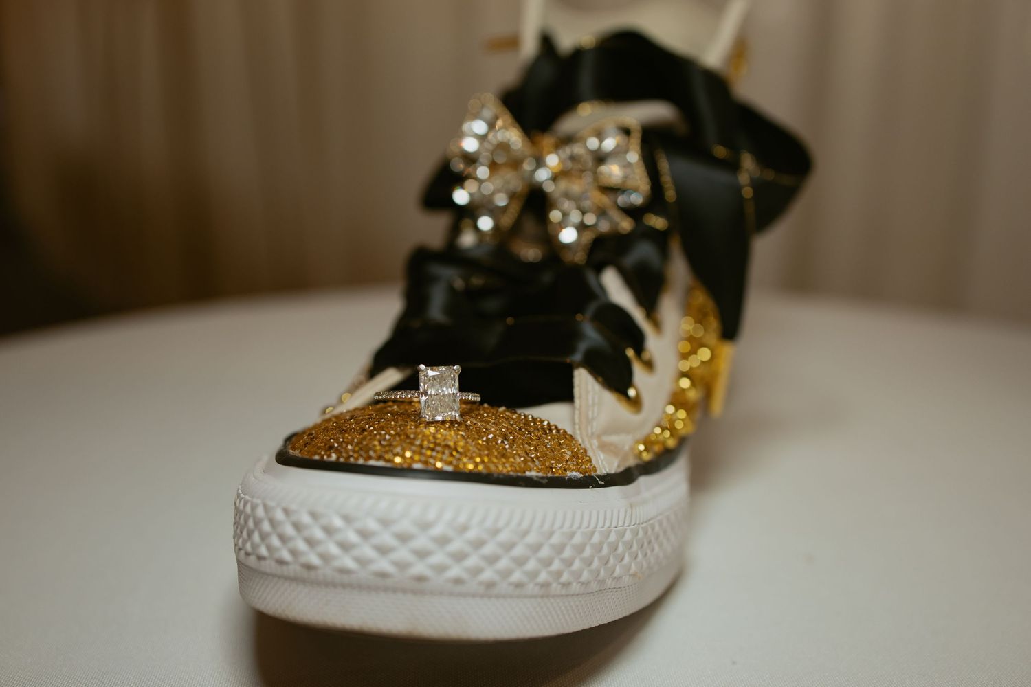 Gold rhinestone Converse sneaker with black bow and diamond ring displayed on white surface.