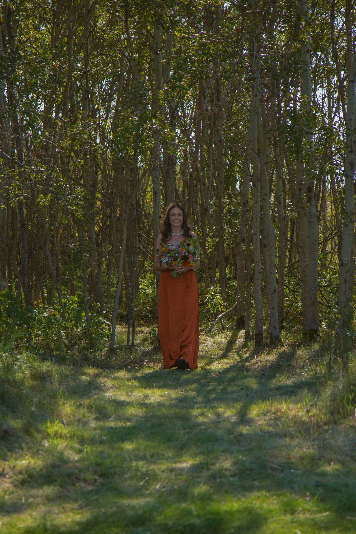 Figure in orange dress standing in a wooded area with dappled sunlight.