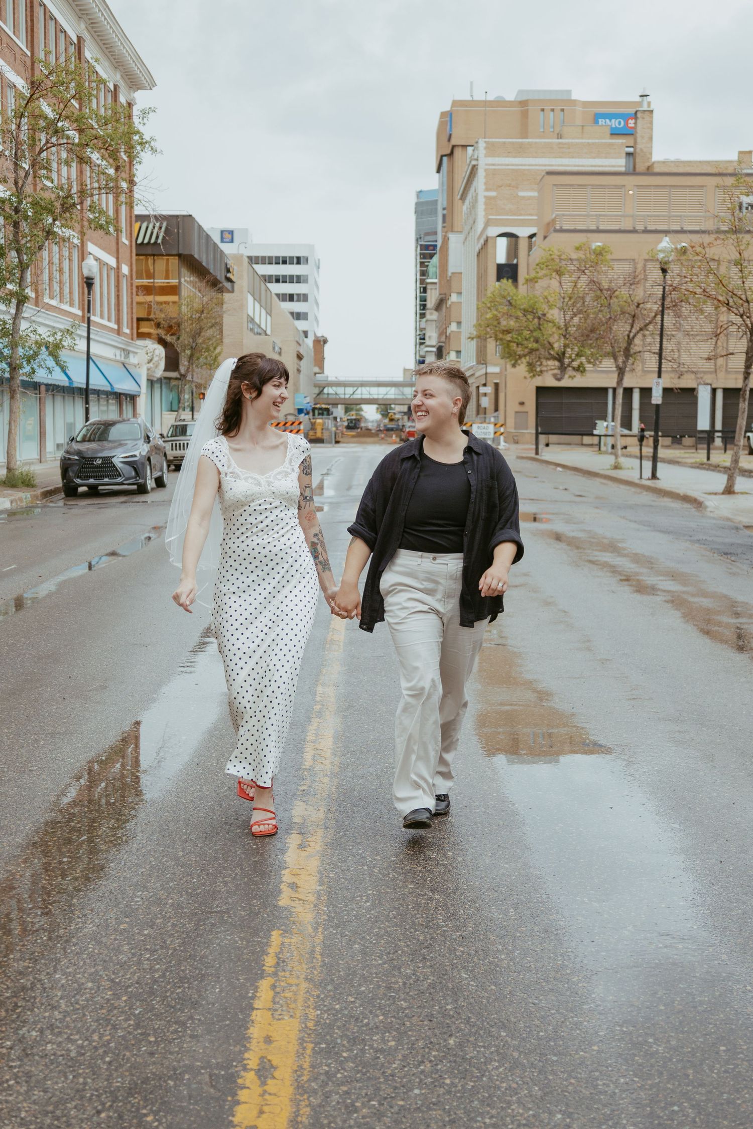 A couple walks together on a wet city street with urban buildings in background.
