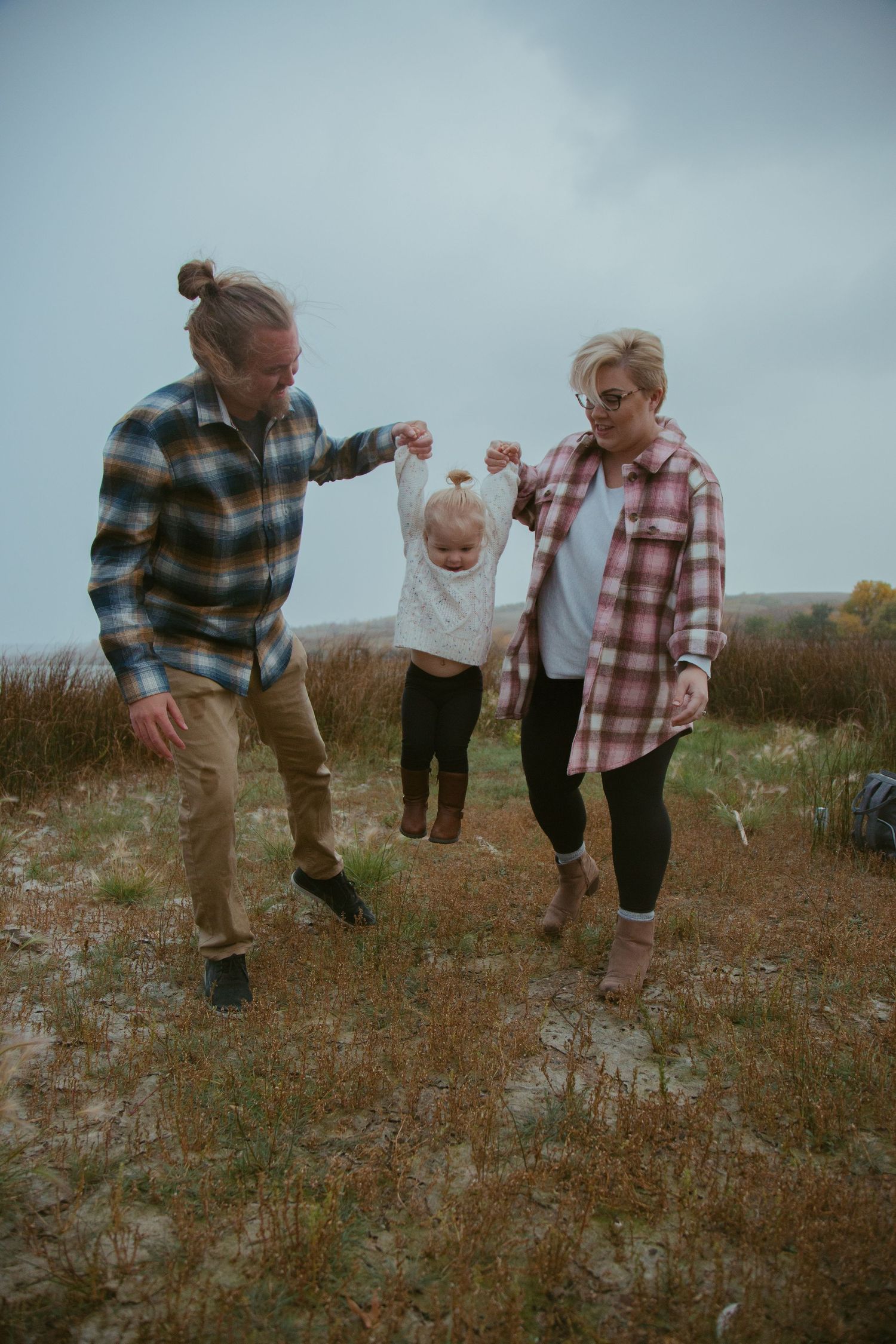Three people in plaid shirts playing and swinging a small child outdoors on an overcast day.