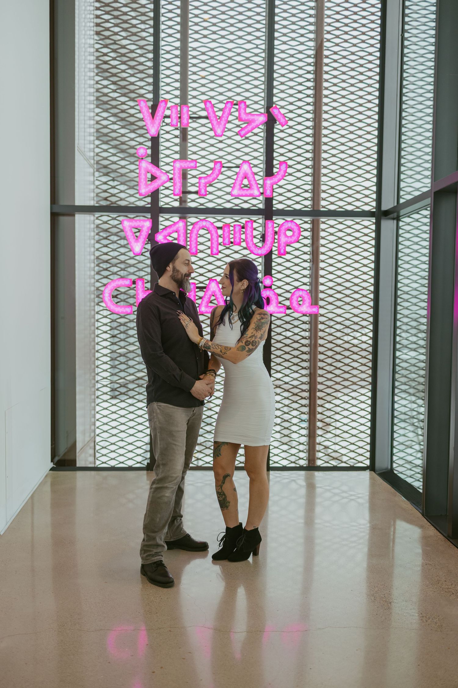 Couple holding hands in front of pink neon text art installation in a modern gallery space.