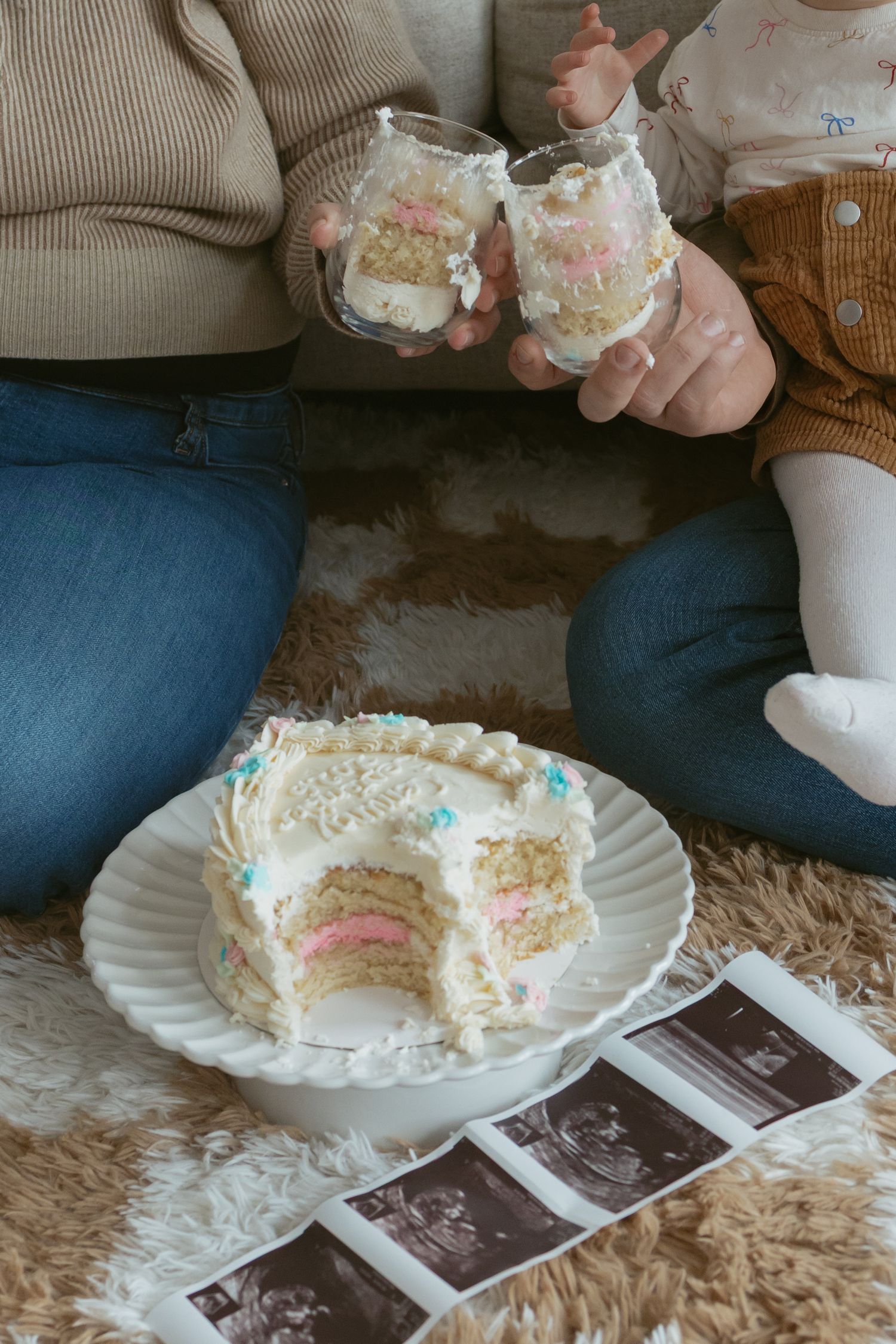 Gender reveal cake with pink layers on plate, ultrasound photos, and two people holding cake jars on a fluffy rug.