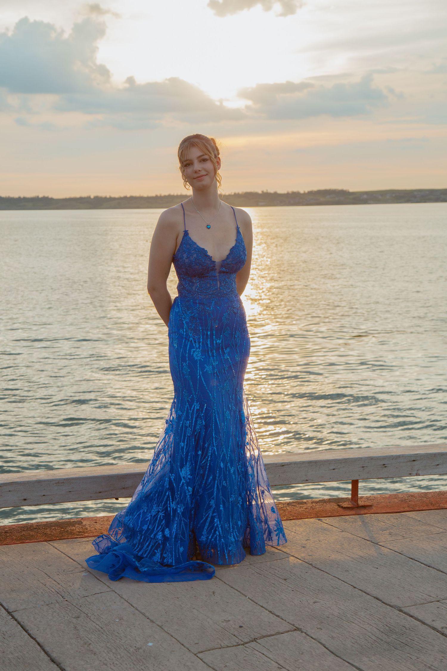 Person in vibrant blue dress sitting on wooden dock at sunset over calm water.