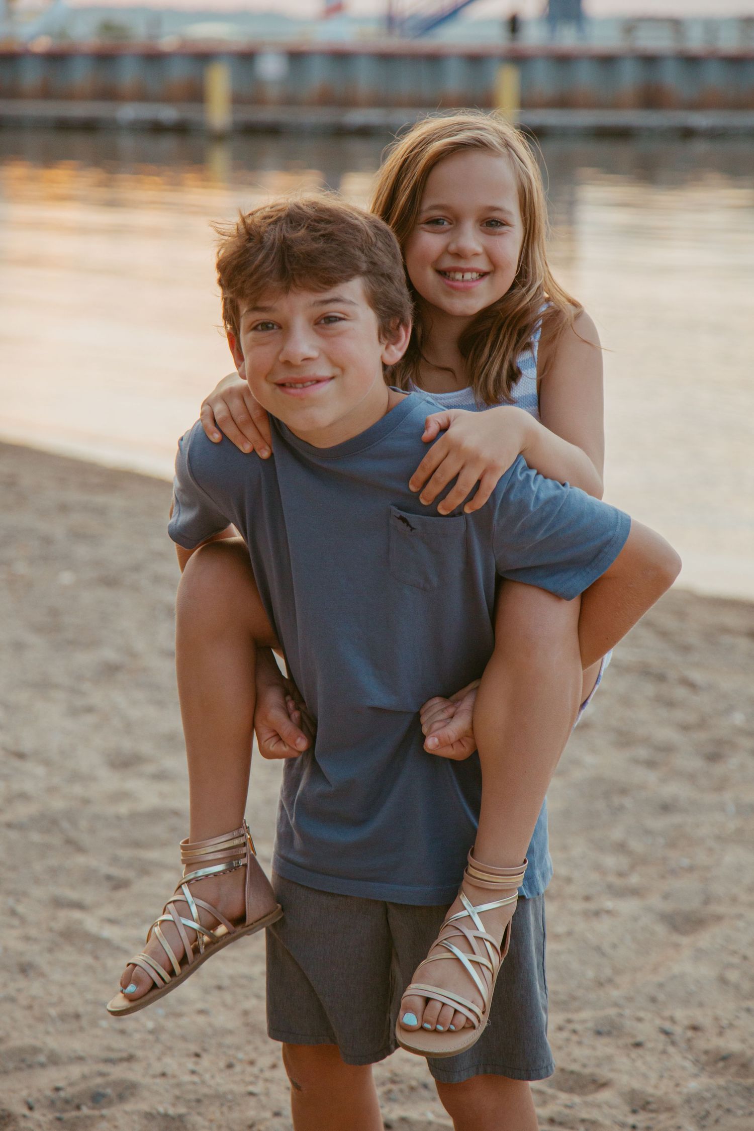 Playful piggyback pose of two children on beach with pier in background at sunset.