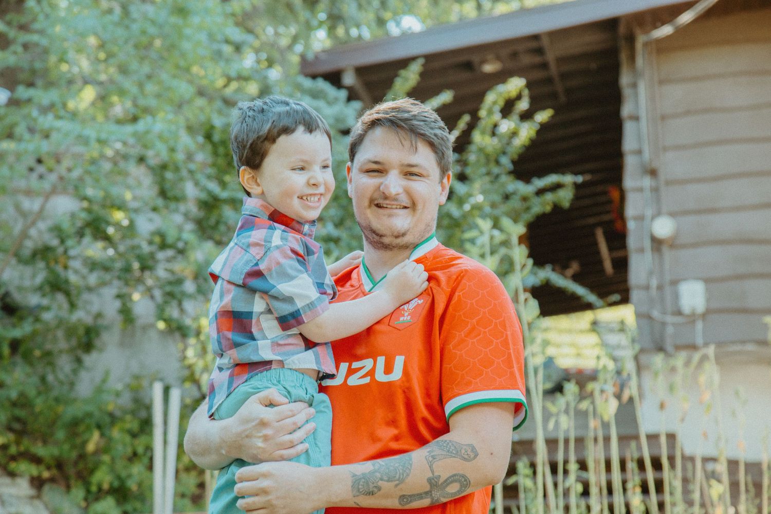 A smiling man in an orange Isuzu shirt holds a young boy in a plaid shirt outdoors near trees and a wooden shed.