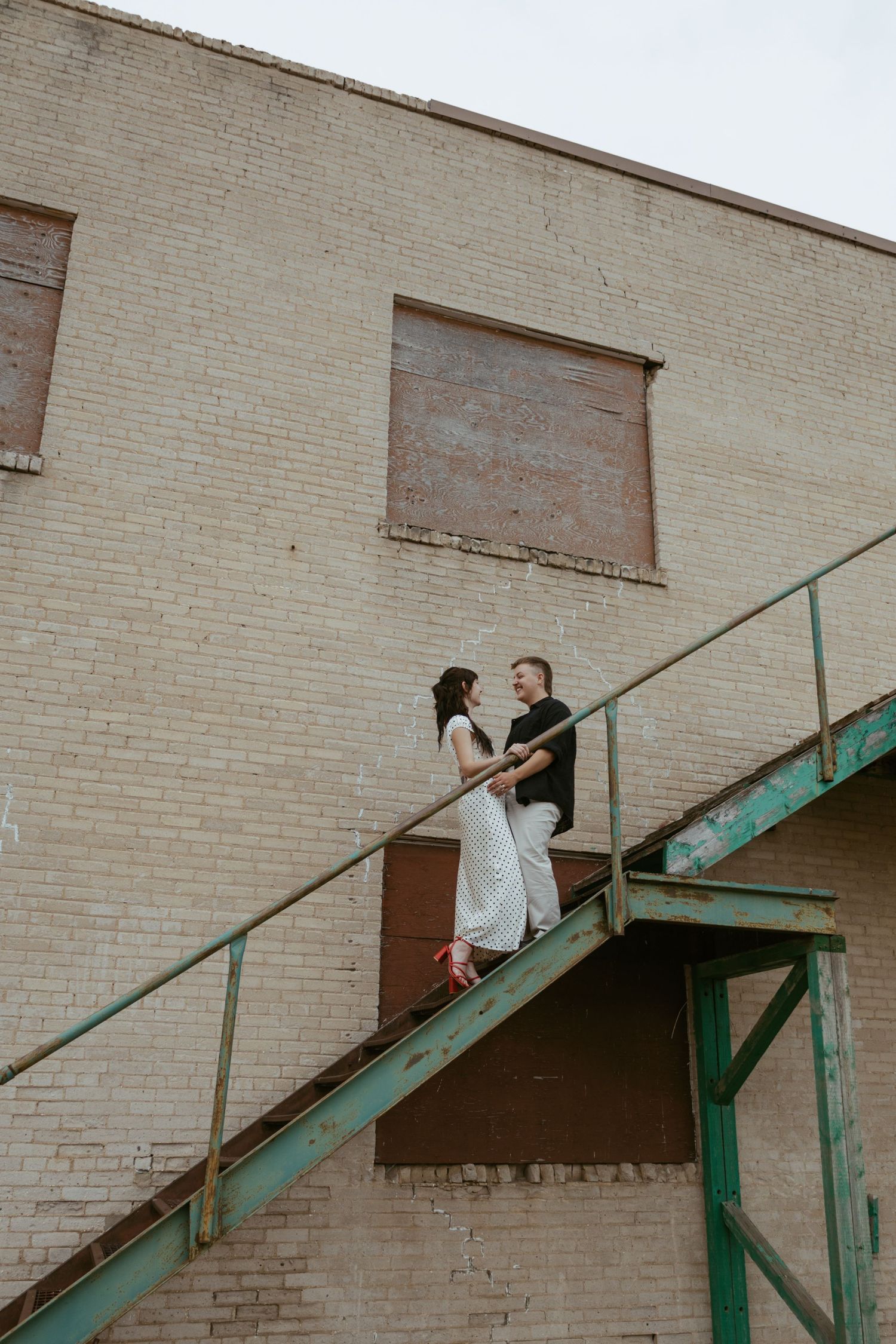 A couple shares a romantic moment on vintage green metal stairs against a brick building.
