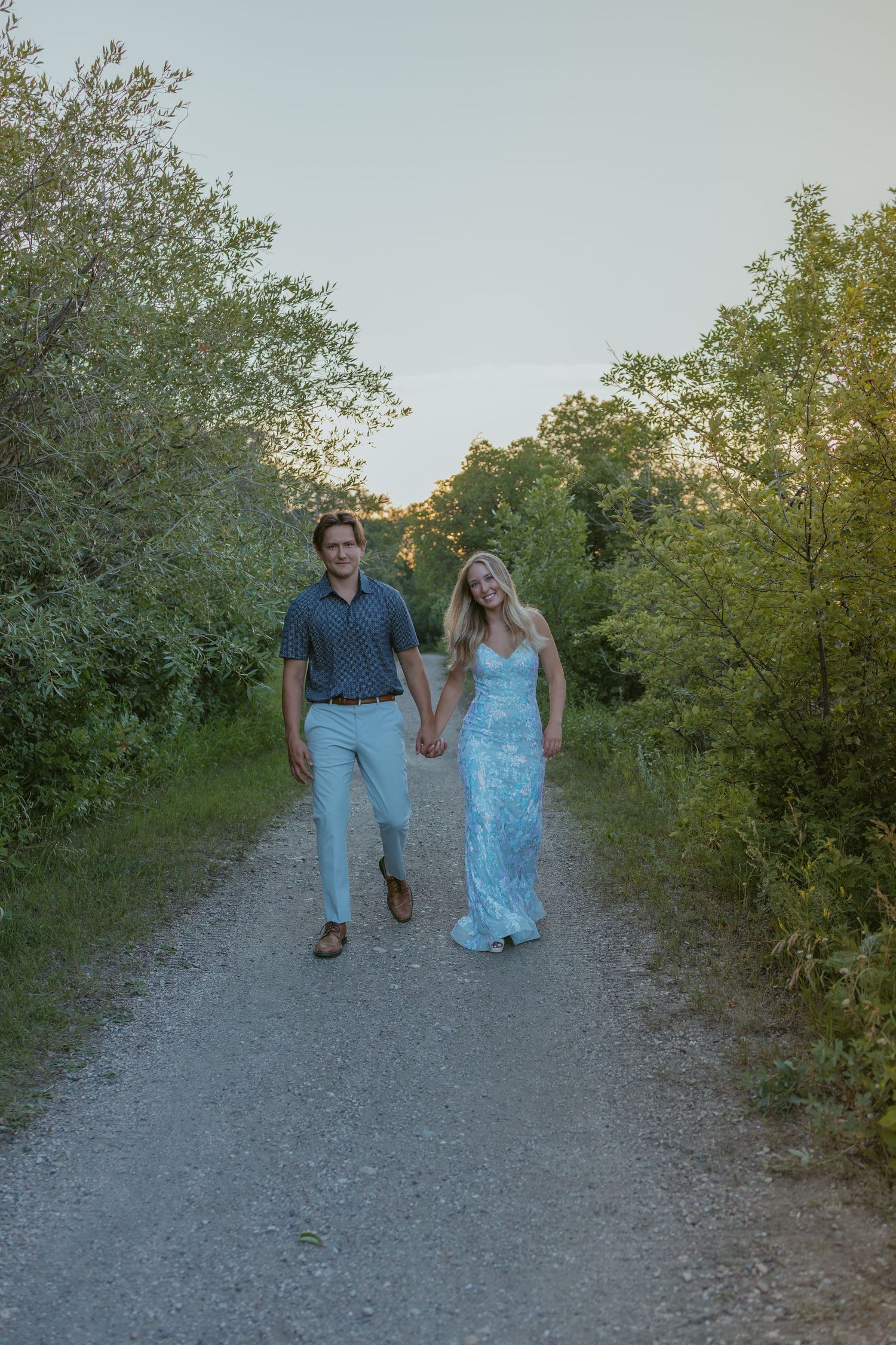 A couple walks together on a gravel path surrounded by green bushes at dusk.