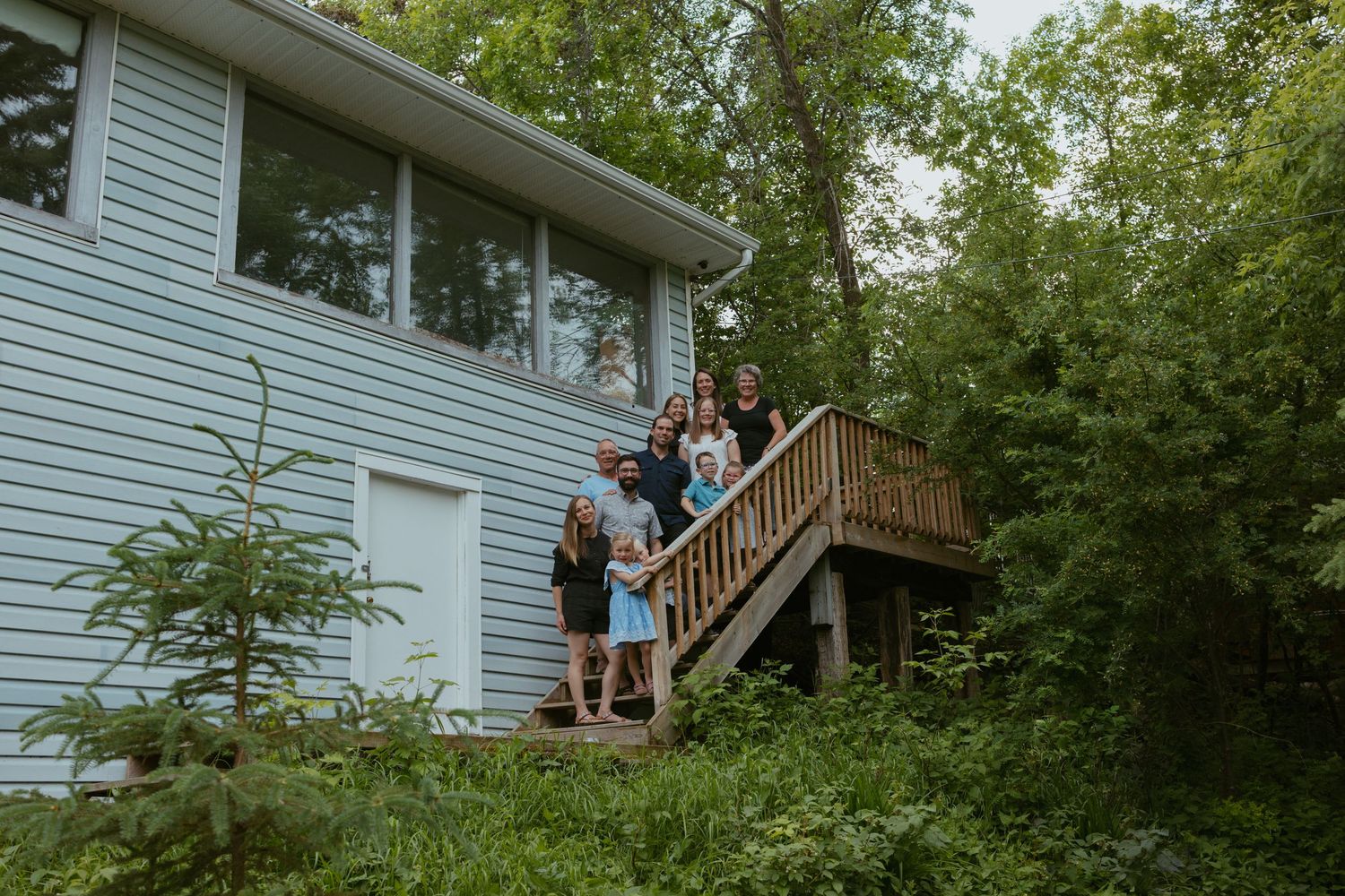 People stand on the deck of a white house surrounded by green trees.